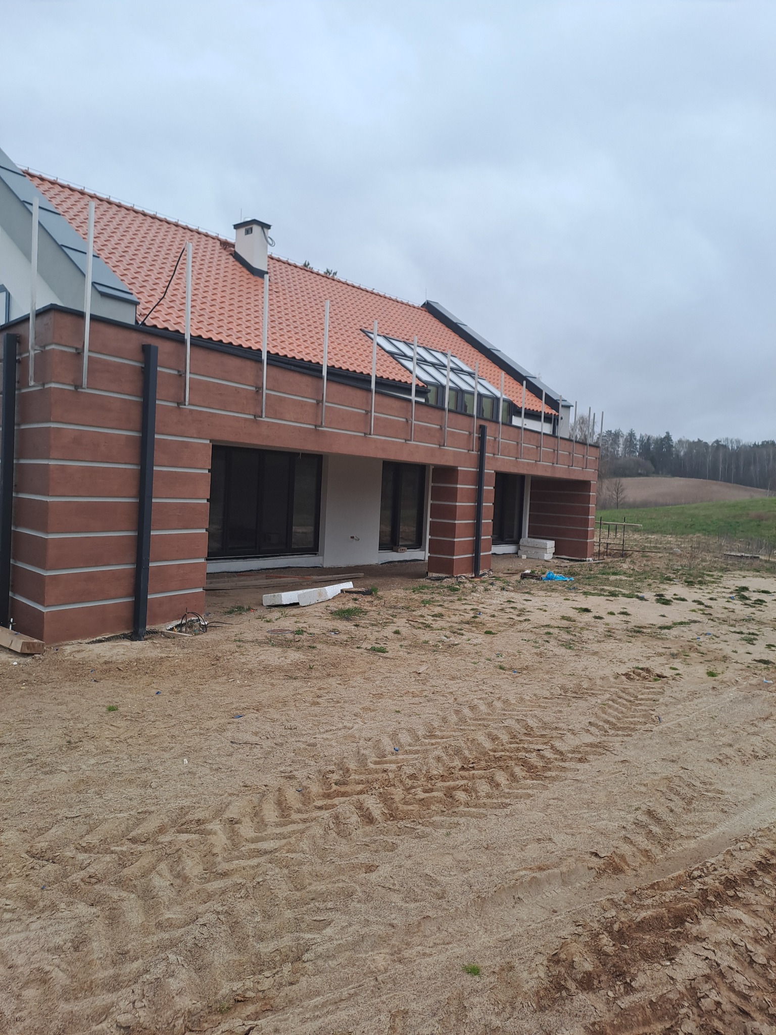 Nowoczesny, dwukondygnacyjny dom z elewacją w odcieniach brązu i beżu, z czerwoną dachówką i oknami dachowymi, widoczny z zewnątrz na tle pochmurnego nieba i pola z wyjeżdżoną ziemią.