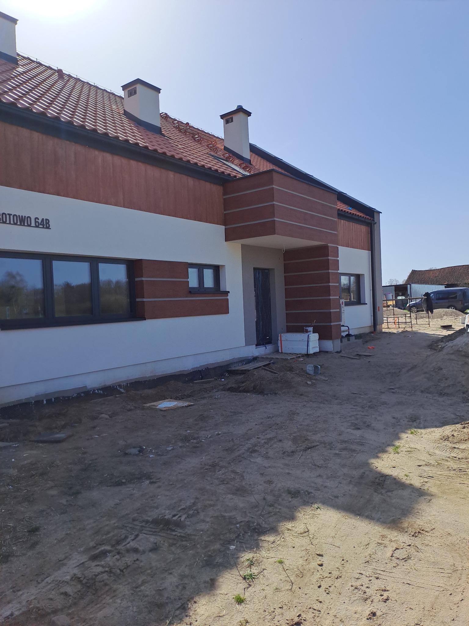 Nowo wybudowany dom jednorodzinny z elementami elewacji wykończonymi w brązowej okładzinie, widoczne styropianowe płyty przy wejściu, teren wokół domu jeszcze niezagospodarowany.