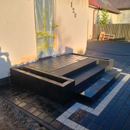 Bruk Proces - Ułożona czarna kostka brukowa przed domem z ciemnymi schodami i donicą, widoczny fragment dekoracyjnej białej kostki, drzwi zabezpieczone folią i taśmą ostrzegawczą.