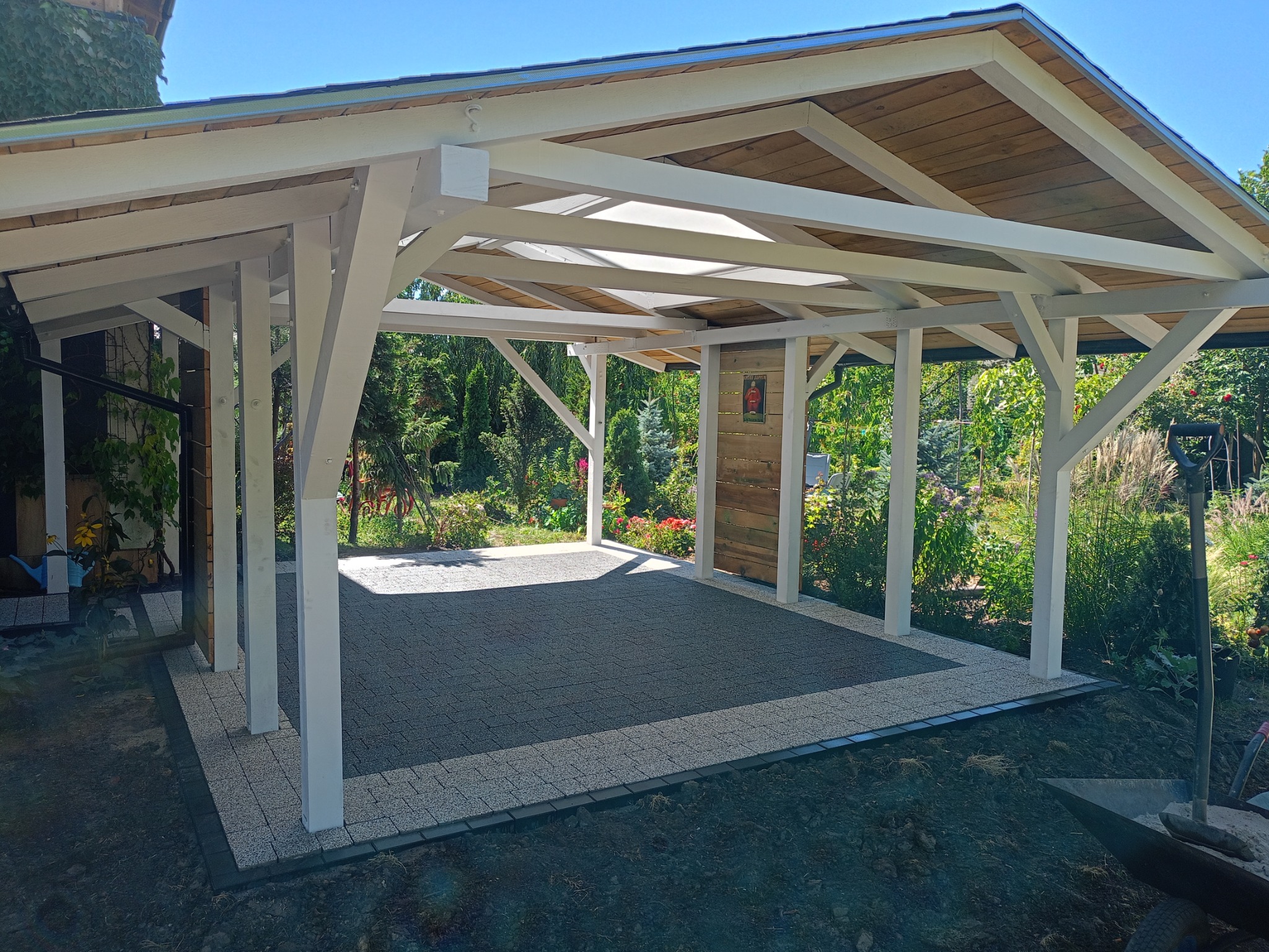 Zadaszona wiata z drewnianą konstrukcją i podłogą wyłożoną szarą kostką brukową, otoczona zielenią ogrodu. Na ścianie wisi dekoracyjny element z motywem muzycznym.