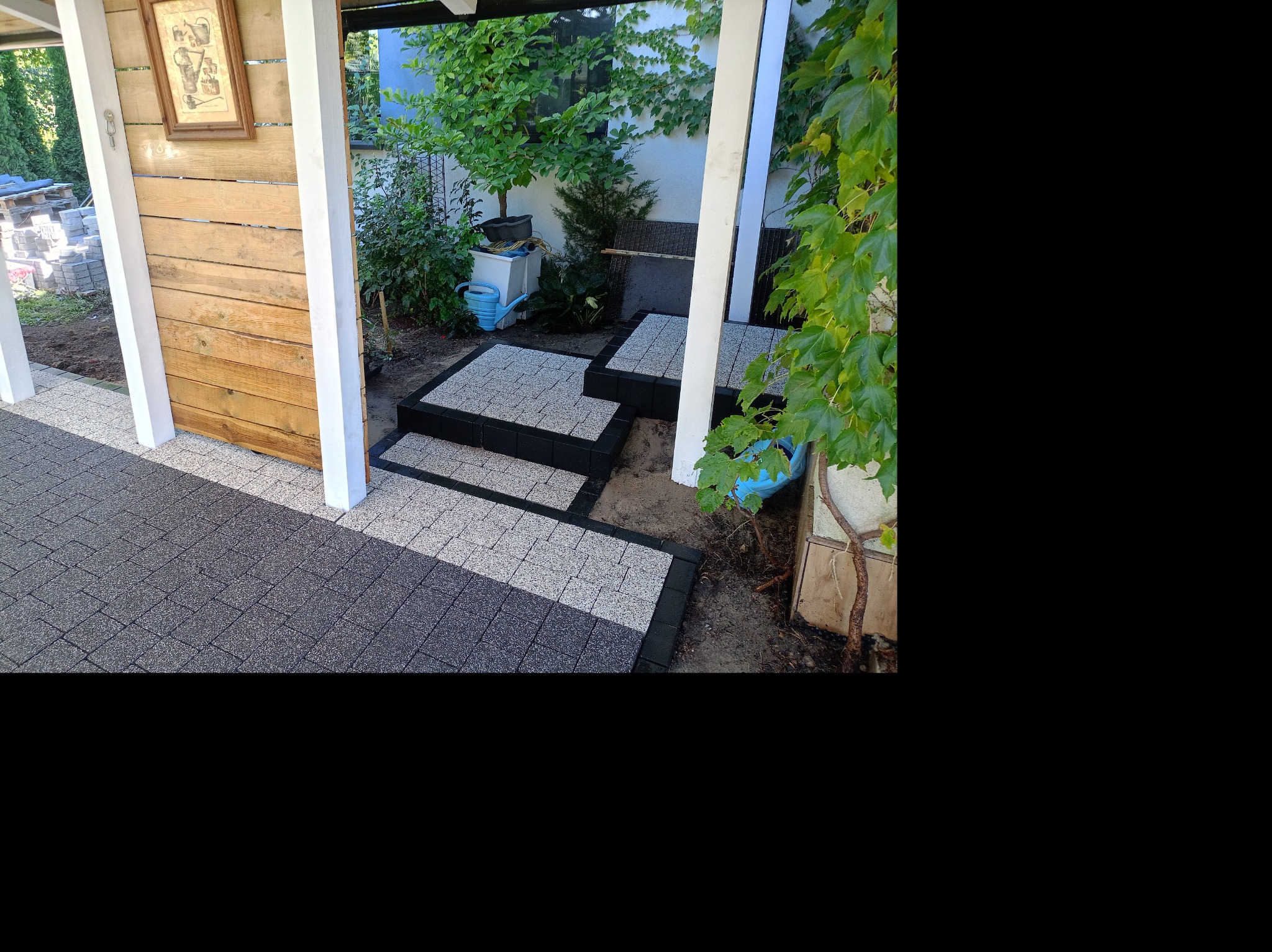 Ułożona nawierzchnia z kostki brukowej w dwóch odcieniach szarości, tworząca ścieżkę i schody w ogrodzie, z drewnianym elementem konstrukcyjnym i roślinnością w tle.