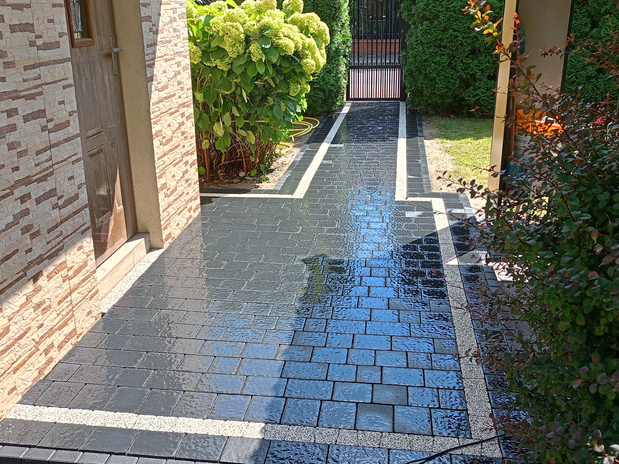 Świeżo ułożona, mokra kostka brukowa w kolorze grafitowym z jasnymi, żwirowymi pasami oddzielającymi, prowadząca od drzwi wejściowych do bramy w tle, otoczona zielenią.