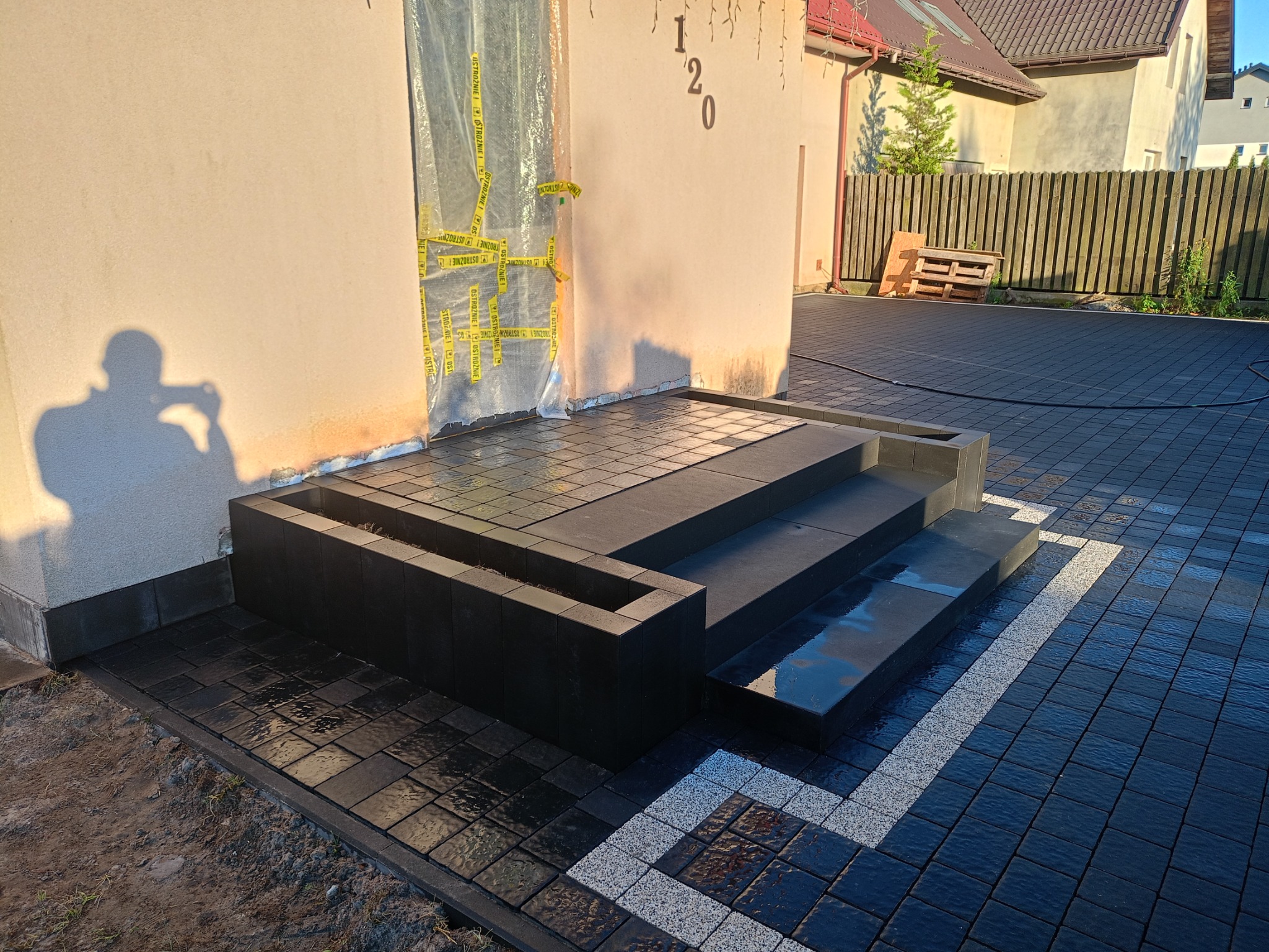 Ułożona czarna kostka brukowa przed domem z ciemnymi schodami i donicą, widoczny fragment dekoracyjnej białej kostki, drzwi zabezpieczone folią i taśmą ostrzegawczą.