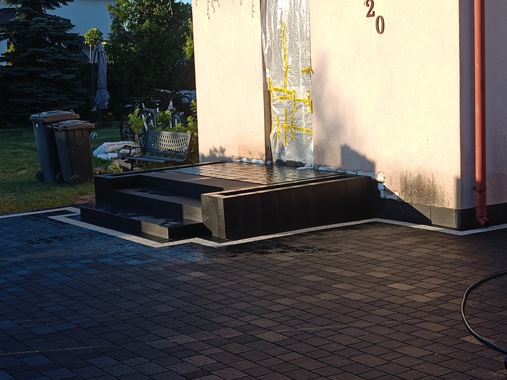 Nowo ułożona, ciemna kostka brukowa przed wejściem do domu z numerem 20, widoczne schody wejściowe i folia zabezpieczająca elewację.