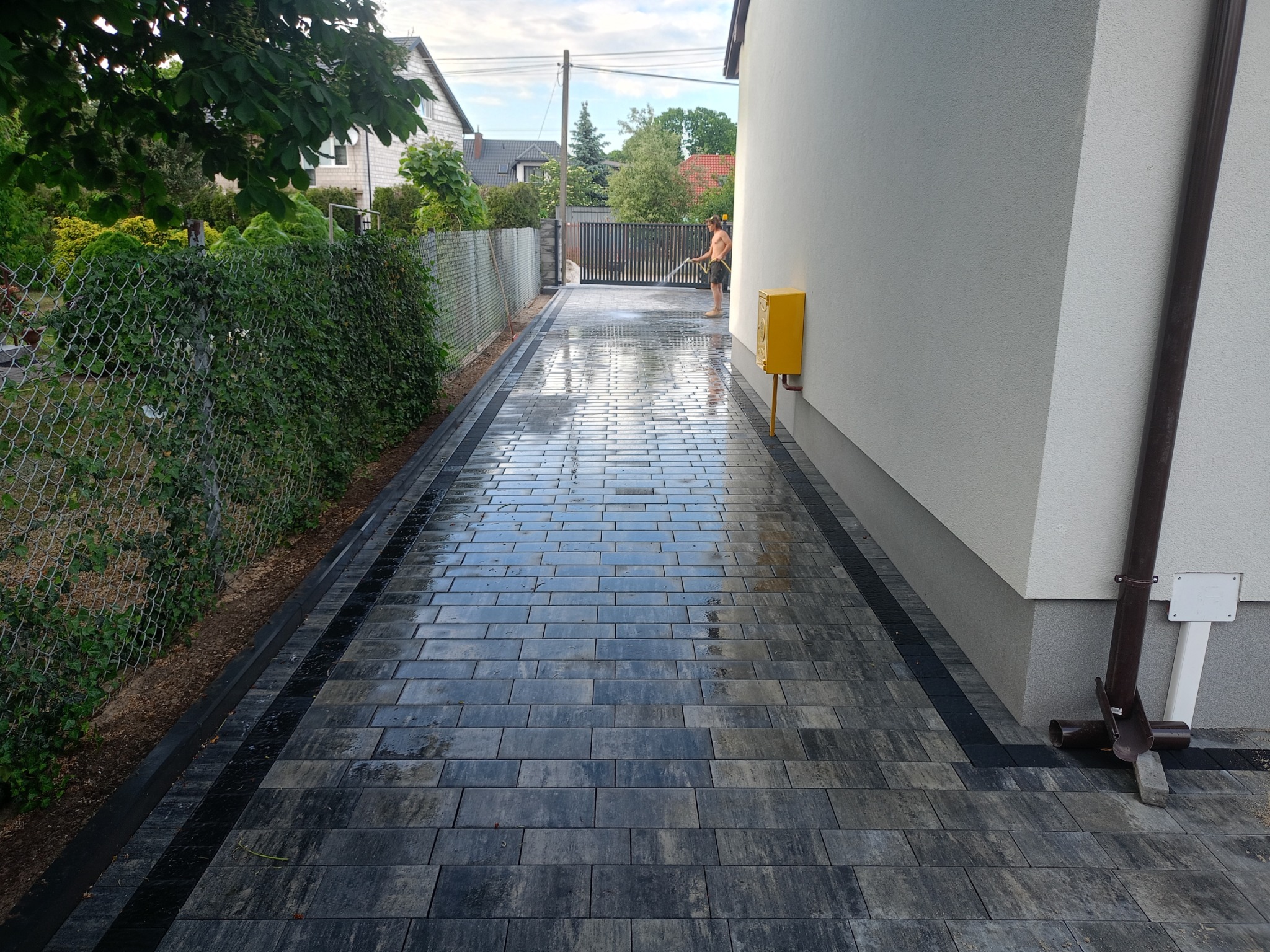 Świeżo ułożona, mokra nawierzchnia z szarej kostki brukowej z czarnymi obrzeżami, myta pod ciśnieniem wzdłuż ogrodzenia z siatki i ściany budynku z żółtą skrzynką gazową.
