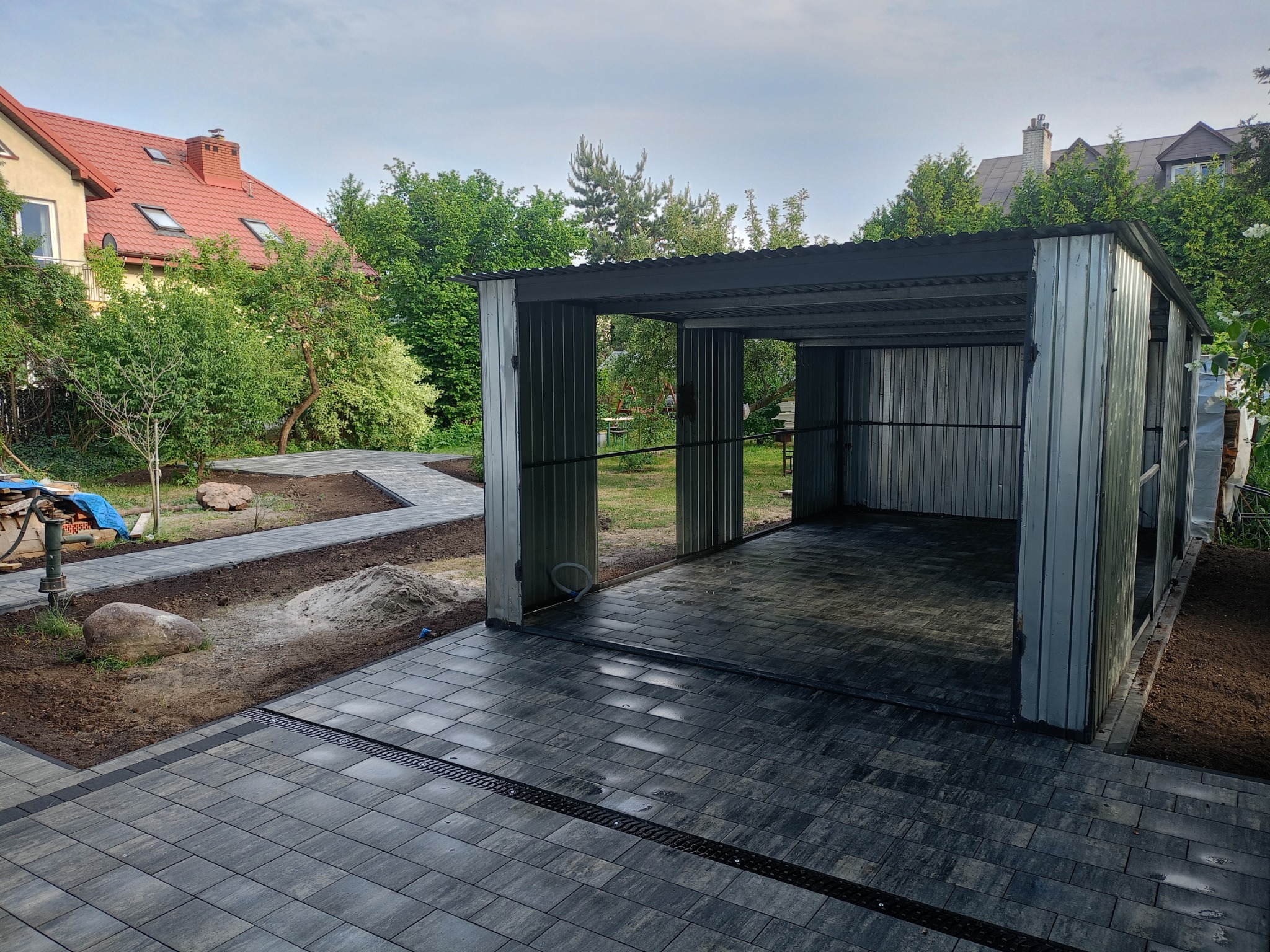 Ułożona kostka brukowa w kolorze grafitowym prowadząca do blaszanej wiaty garażowej z widocznym odpływem liniowym i elementami ogrodu w tle.
