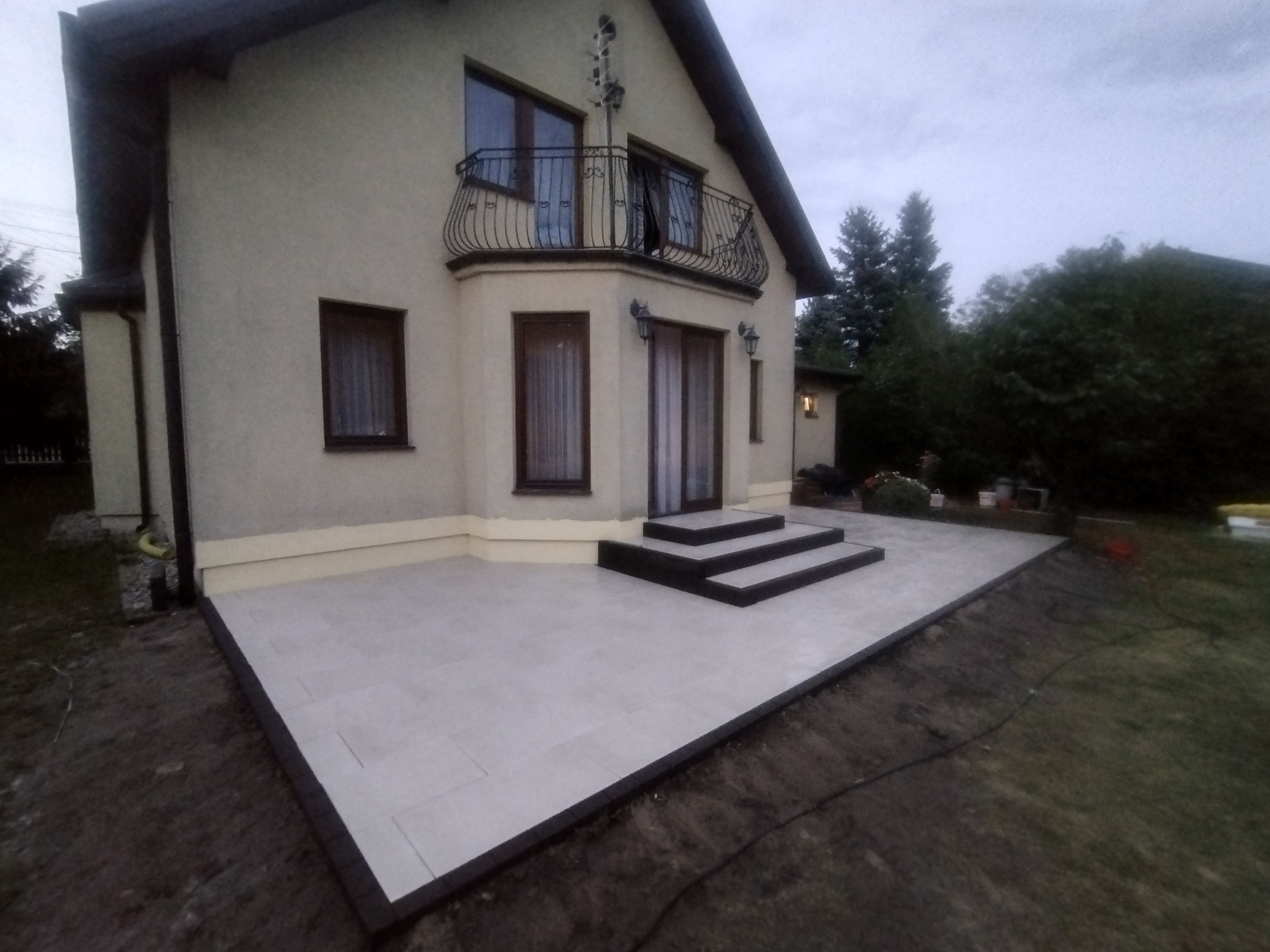Nowoczesny taras wyłożony jasnymi płytami z ciemną obwódką, prowadzący do domu z balkonem, widok z ogrodu.