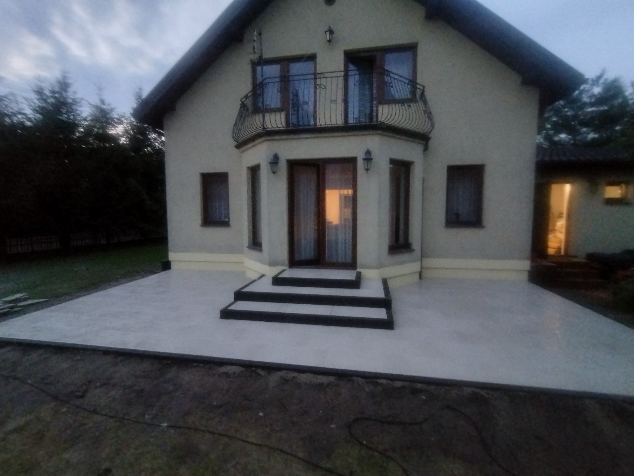 Jasnoszara powierzchnia tarasu przed domem z trzema schodami z ciemnymi obrzeżami, widok na dom z balkonem i oknami w pochmurny dzień.