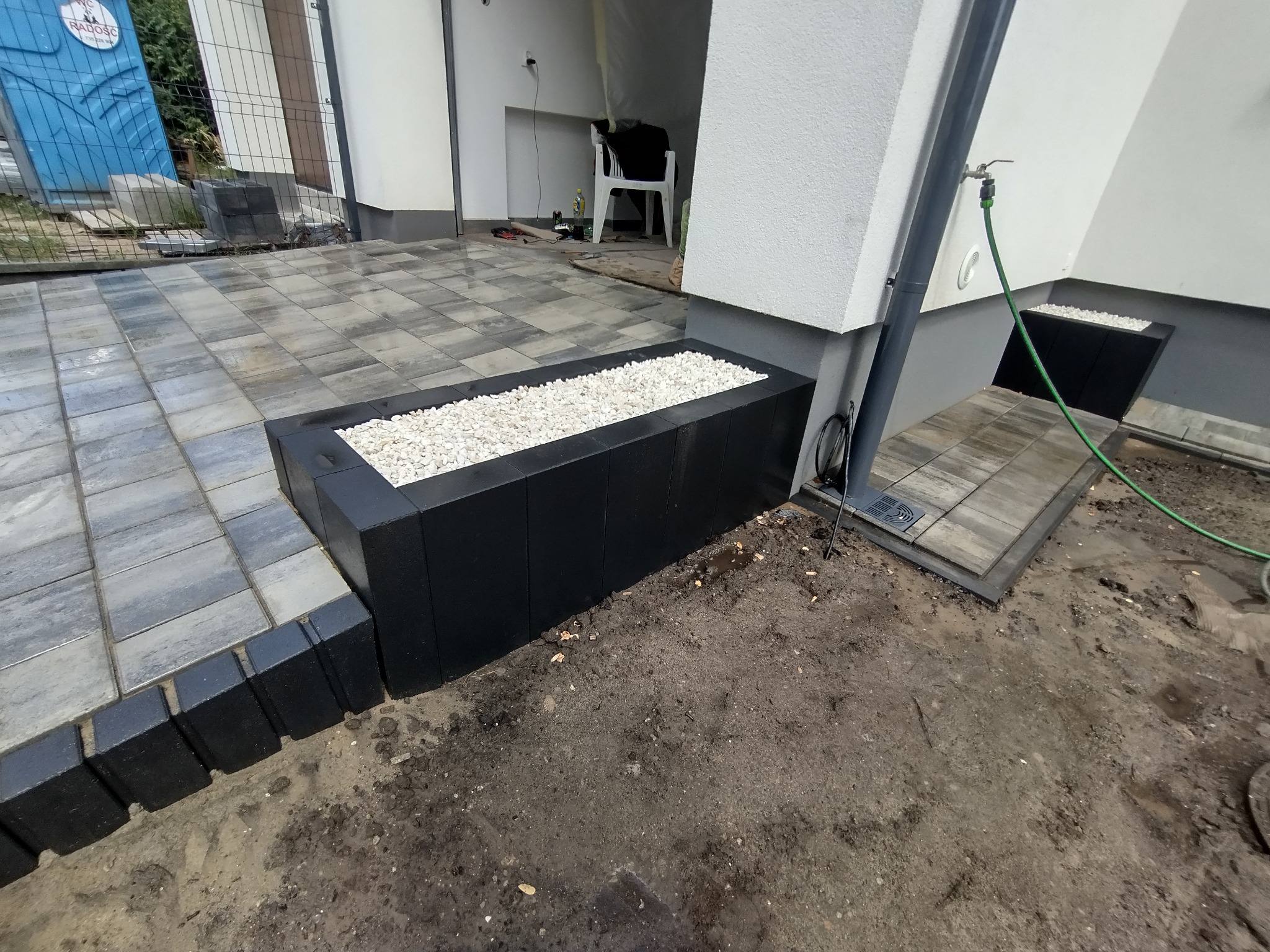 Ułożona szara kostka brukowa z obrzeżami, czarna donica z białymi kamykami, kran z wężem ogrodowym przy ścianie domu.