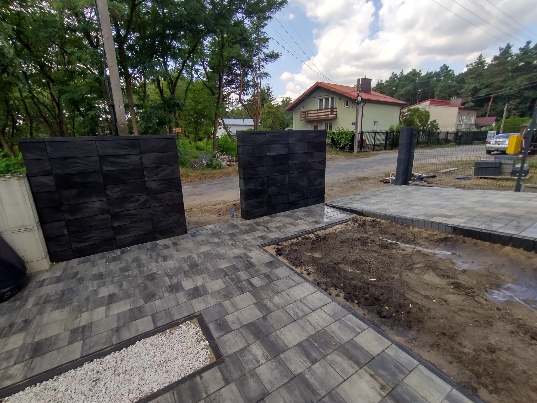 Nowo ułożona szara kostka brukowa na podjeździe z czarnymi, modułowymi słupkami ogrodzeniowymi; fragment przygotowanej ziemi pod dalszą część podjazdu.