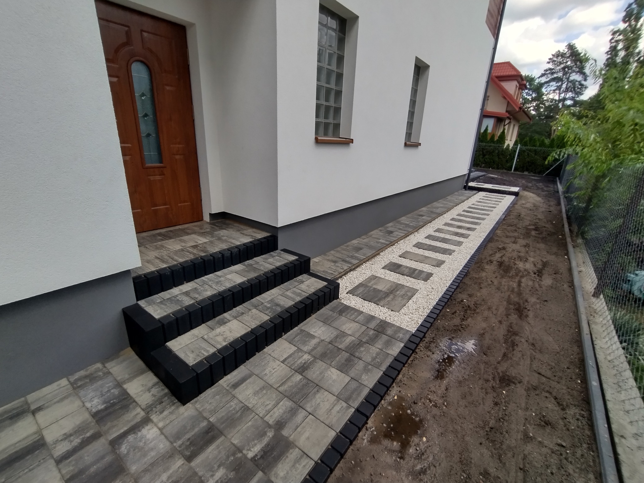Nowoczesne schody wejściowe i ścieżka z kostki brukowej w odcieniach szarości, obramowane czarnym obrzeżem, prowadzące do białego domu z drewnianymi oknami.