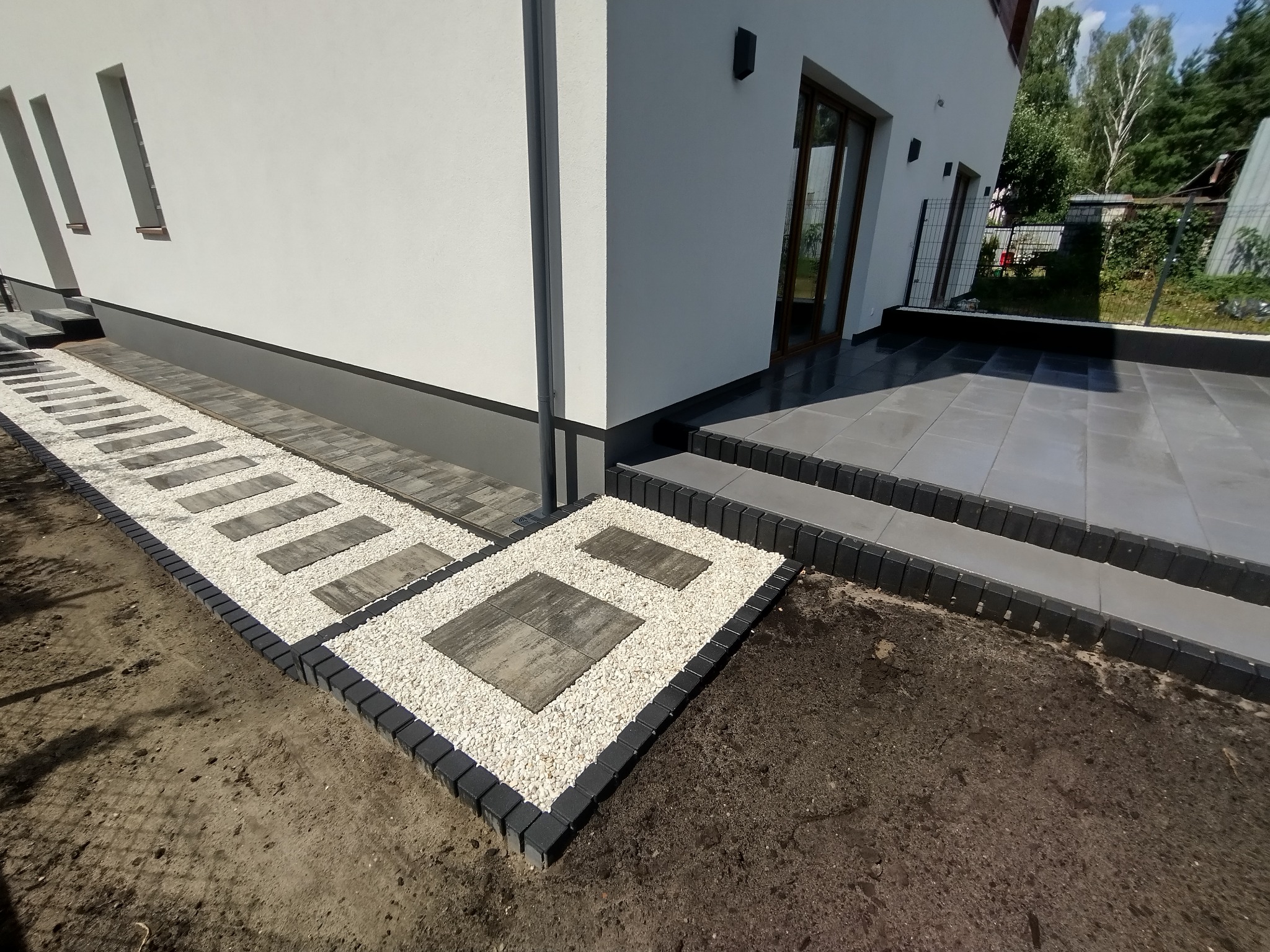 Nowoczesna ścieżka z szarych płyt i białego grysu, obrzeża z ciemnej kostki, prowadząca do domu z szarą elewacją i tarasem z szarych płyt.