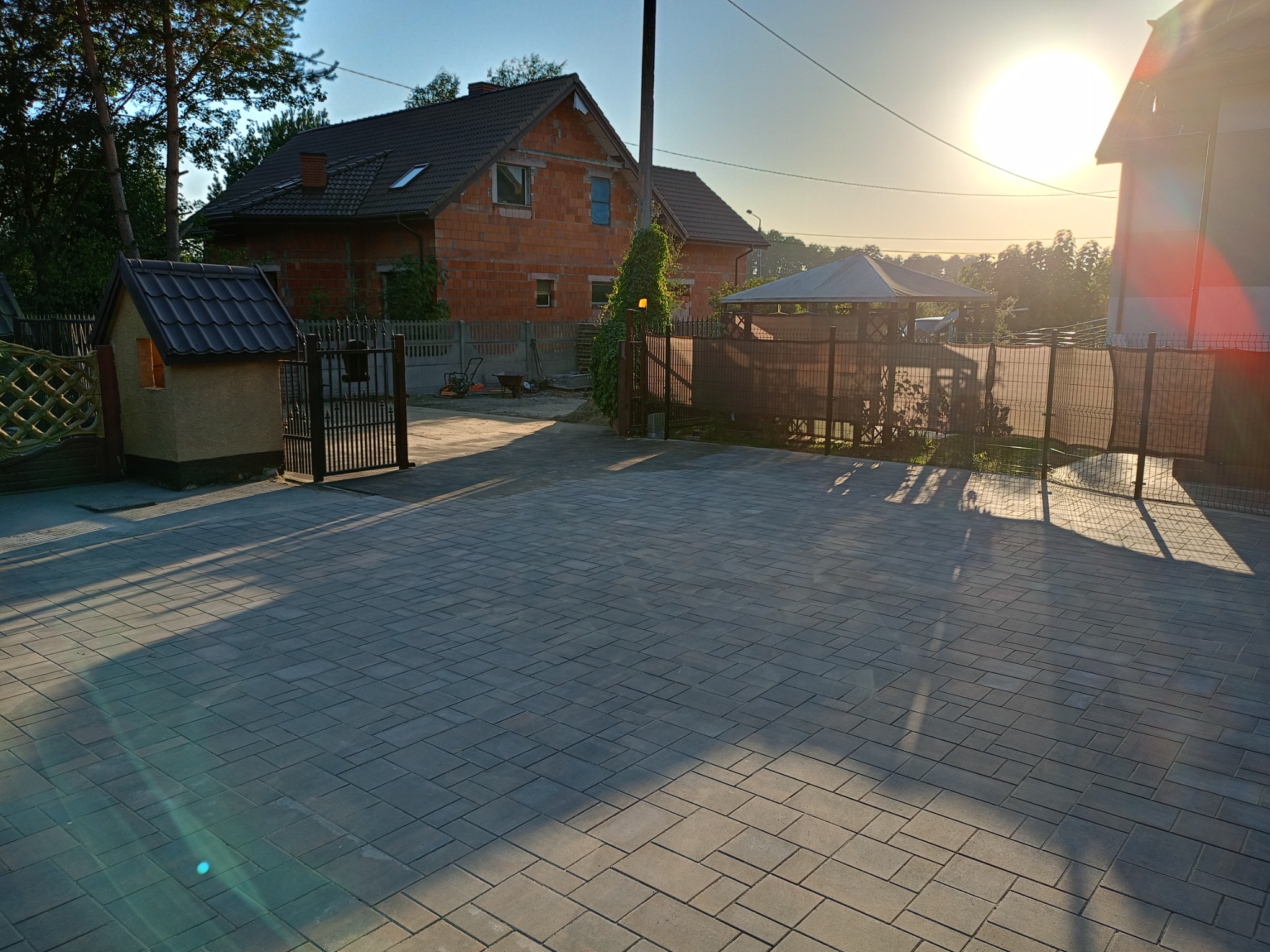Nowo ułożona, szara kostka brukowa na podjeździe przed domem w trakcie wschodu słońca, z widocznym domkiem dla dzieci i altaną w tle.