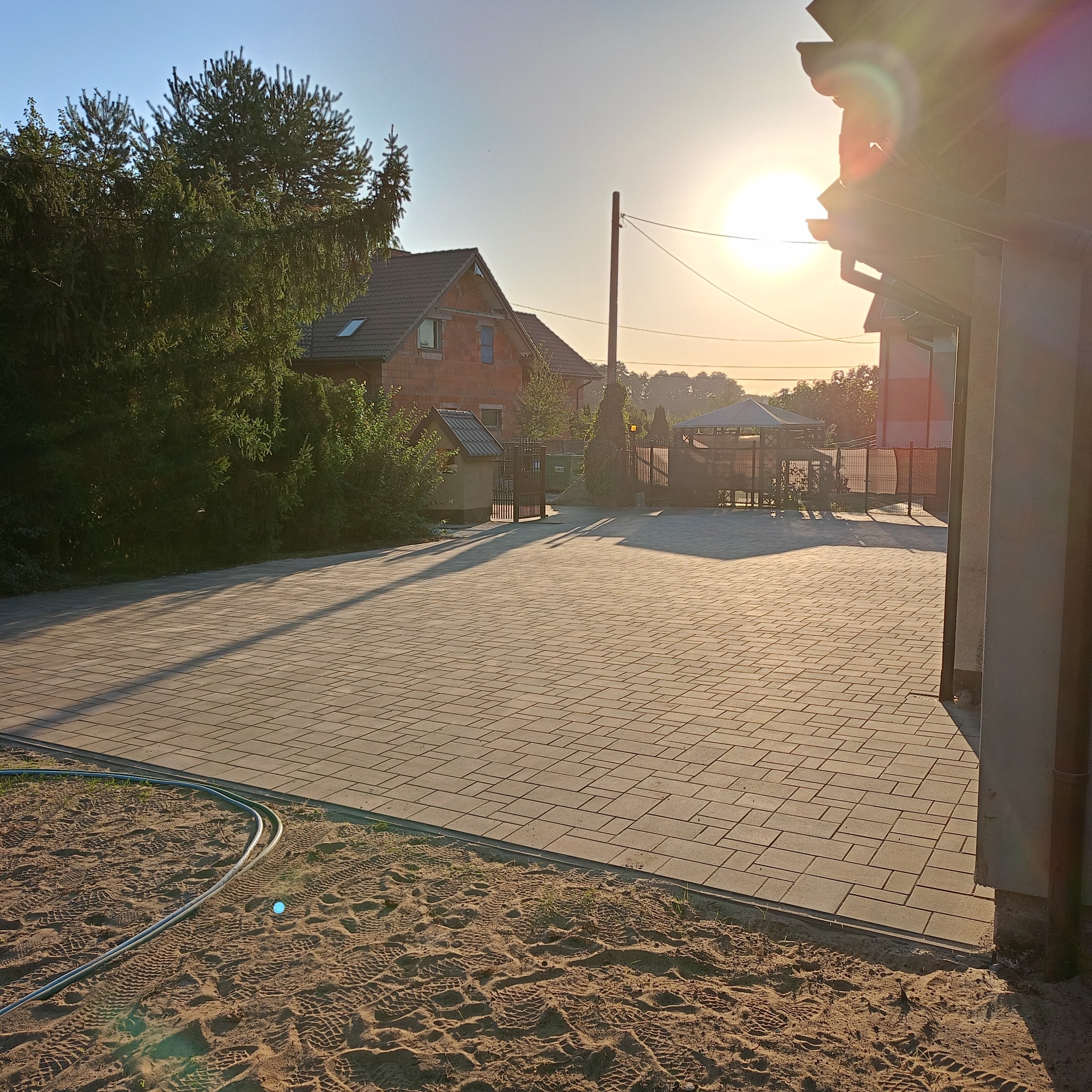 Szeroki podjazd z jasnoszarej kostki brukowej ułożonej we wzór cegły, z widocznym piaskiem na obrzeżach i wężem ogrodowym, w tle dom z czerwonej cegły w promieniach zachodzącego słońca.
