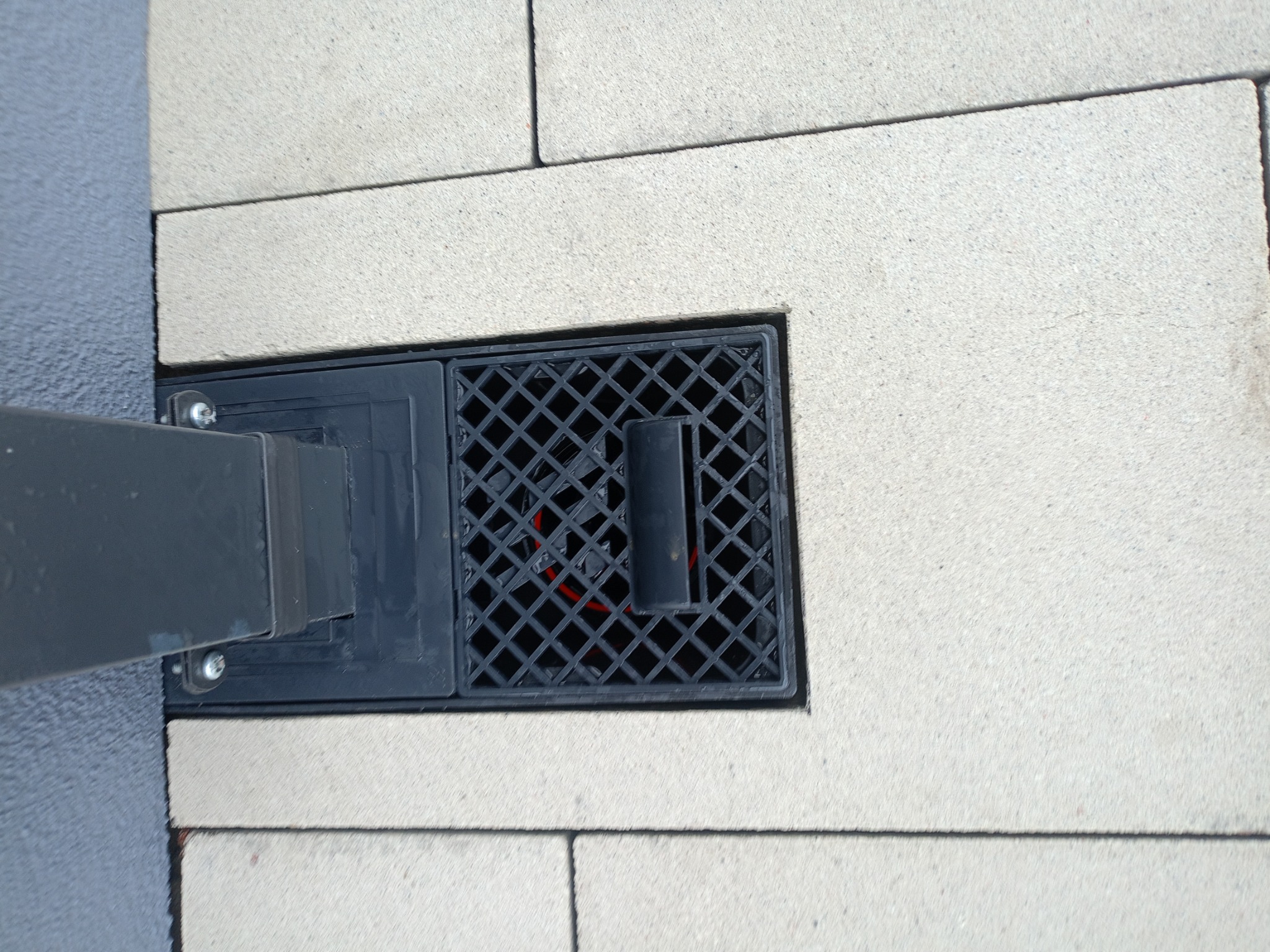 Szary odpływ liniowy wkomponowany w jasną kostkę brukową, widoczne elementy mocujące do słupka.