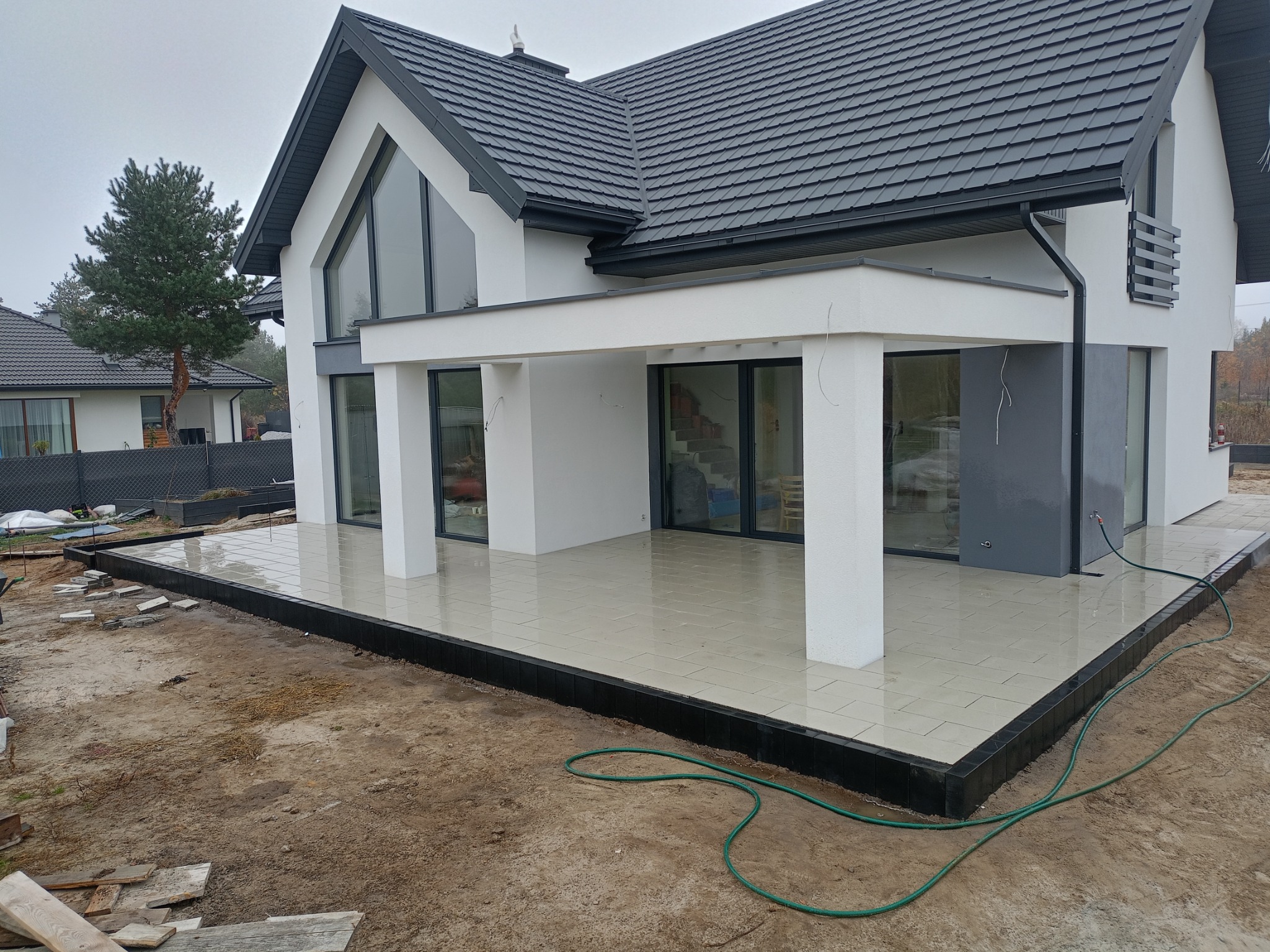 Nowoczesny dom z wyłożonym jasnymi płytami tarasem obramowanym czarnym krawężnikiem, widoczny zielony wąż ogrodowy.