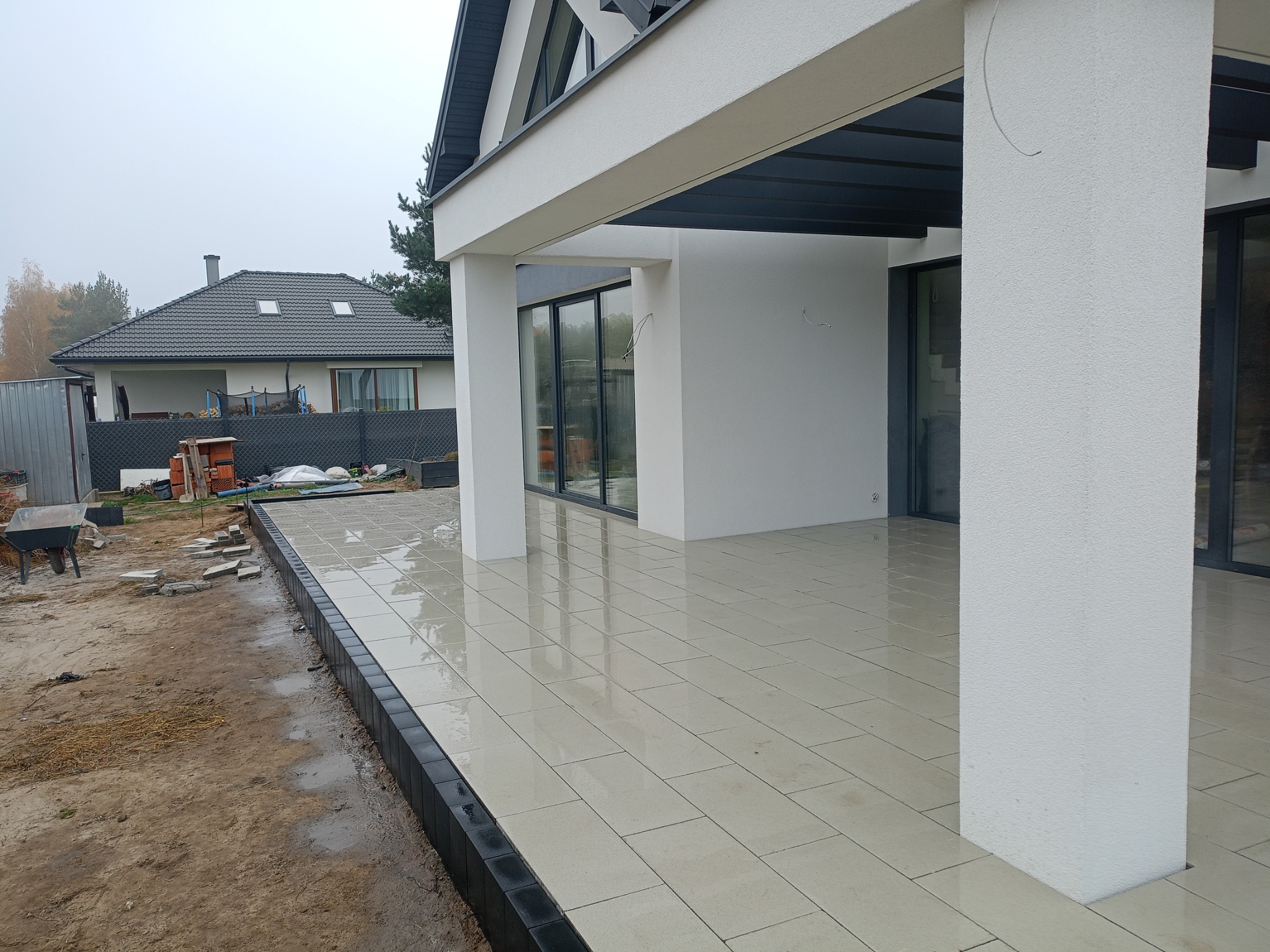 Jasne, prostokątne płyty tarasowe ułożone na zewnątrz nowoczesnego domu z białymi kolumnami, czarną obwódką i widoczną konstrukcją dachu.