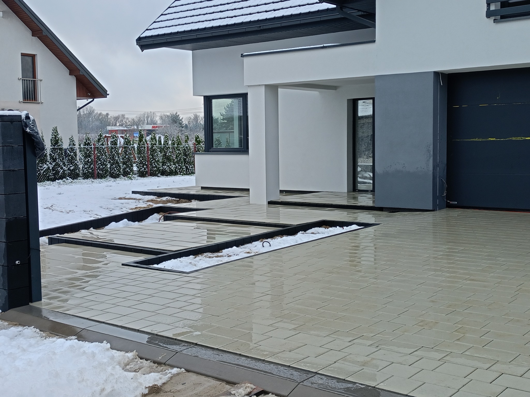 Nowoczesna posesja z jasnoszarą kostką brukową ułożoną w regularny wzór, z czarnymi obrzeżami i śniegiem w szczelinach, na tle minimalistycznego domu z ciemną bramą garażową.