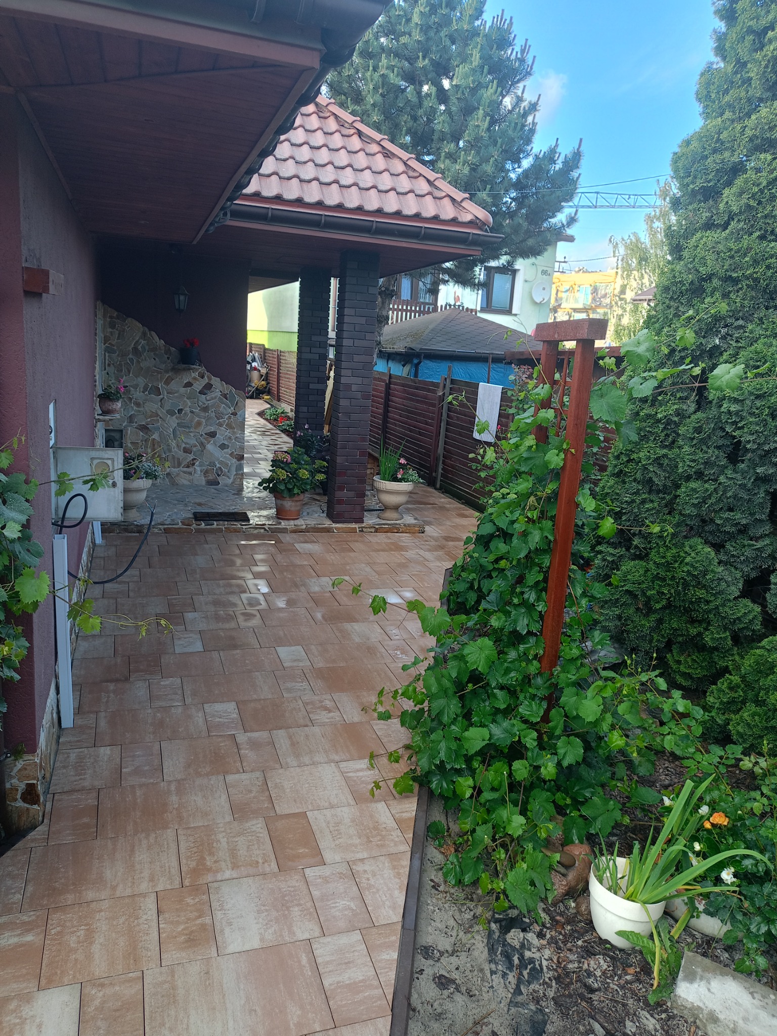 Ułożona, brązowa kostka brukowa prowadząca do domu z kamienną elewacją i kolumną z cegły. Widoczne rośliny i elementy dekoracyjne.