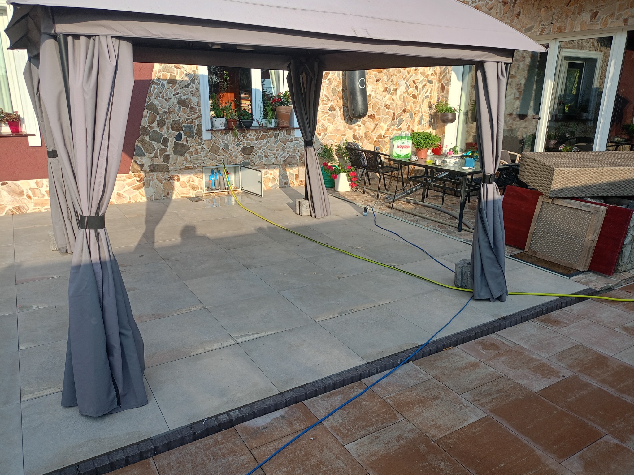 Jasnoszare płyty tarasowe pod altaną, oddzielone ciemną obwódką od brązowej kostki. Kamienna fasada domu w tle, rośliny w donicach na parapecie.