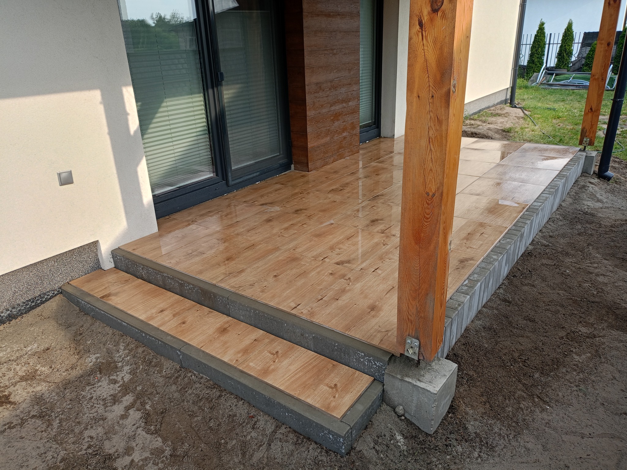 Wykończony taras z płytkami imitującymi drewno, z dwoma stopniami i drewnianą kolumną. Widoczne obrzeża z kostki brukowej i fragment trawnika w tle.