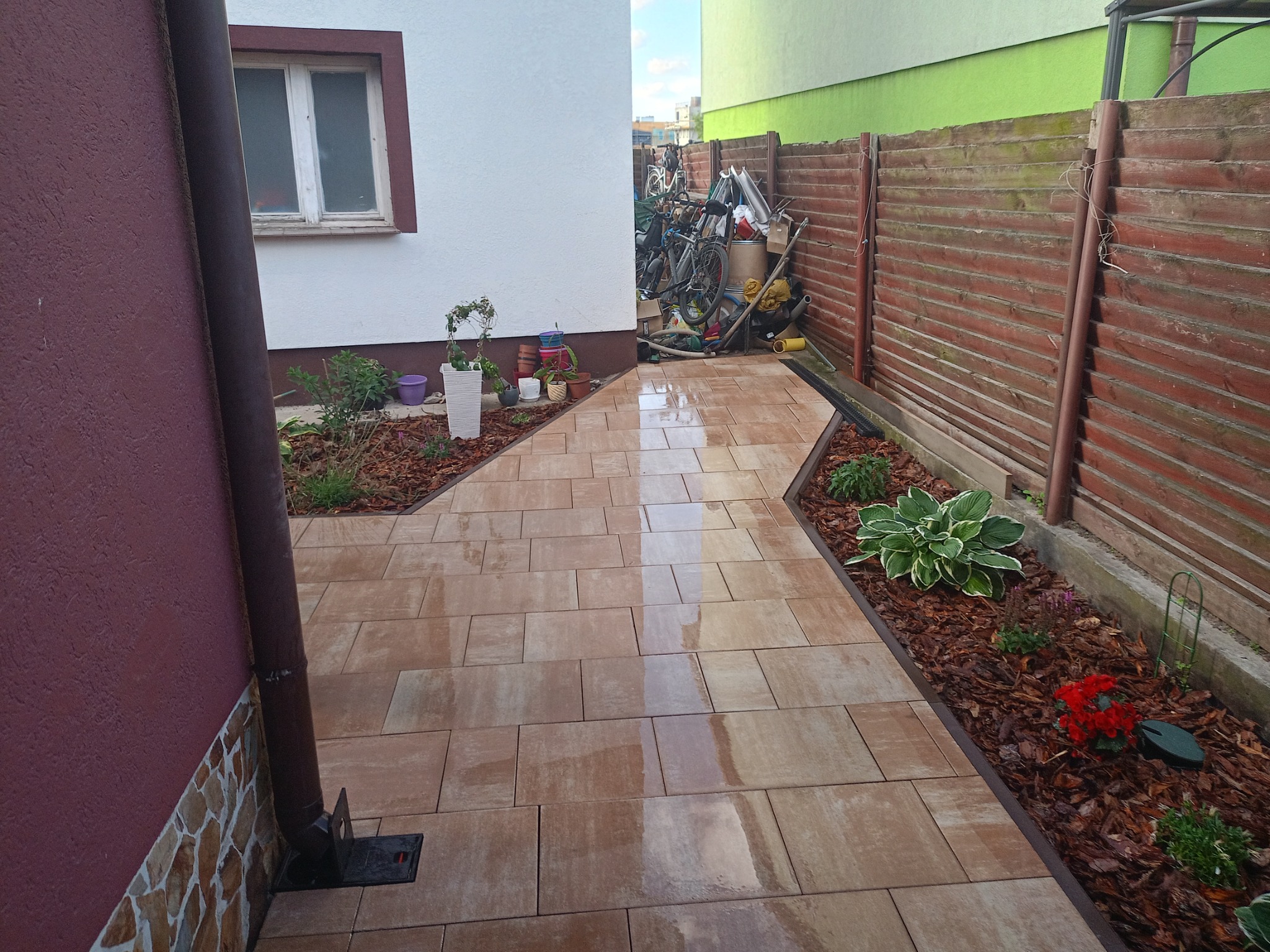 Ułożona, beżowa kostka brukowa tworzy ścieżkę wzdłuż domu z brązową elewacją i drewnianego płotu. Rabaty z korą i roślinami po bokach. W tle rowery i akcesoria.