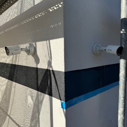 JS-Elektro - Dwie kamery monitoringu Hikvision zamontowane na elewacji budynku z widocznymi cieniami rusztowania i fragmentami malowania. Jasne światło słoneczne.