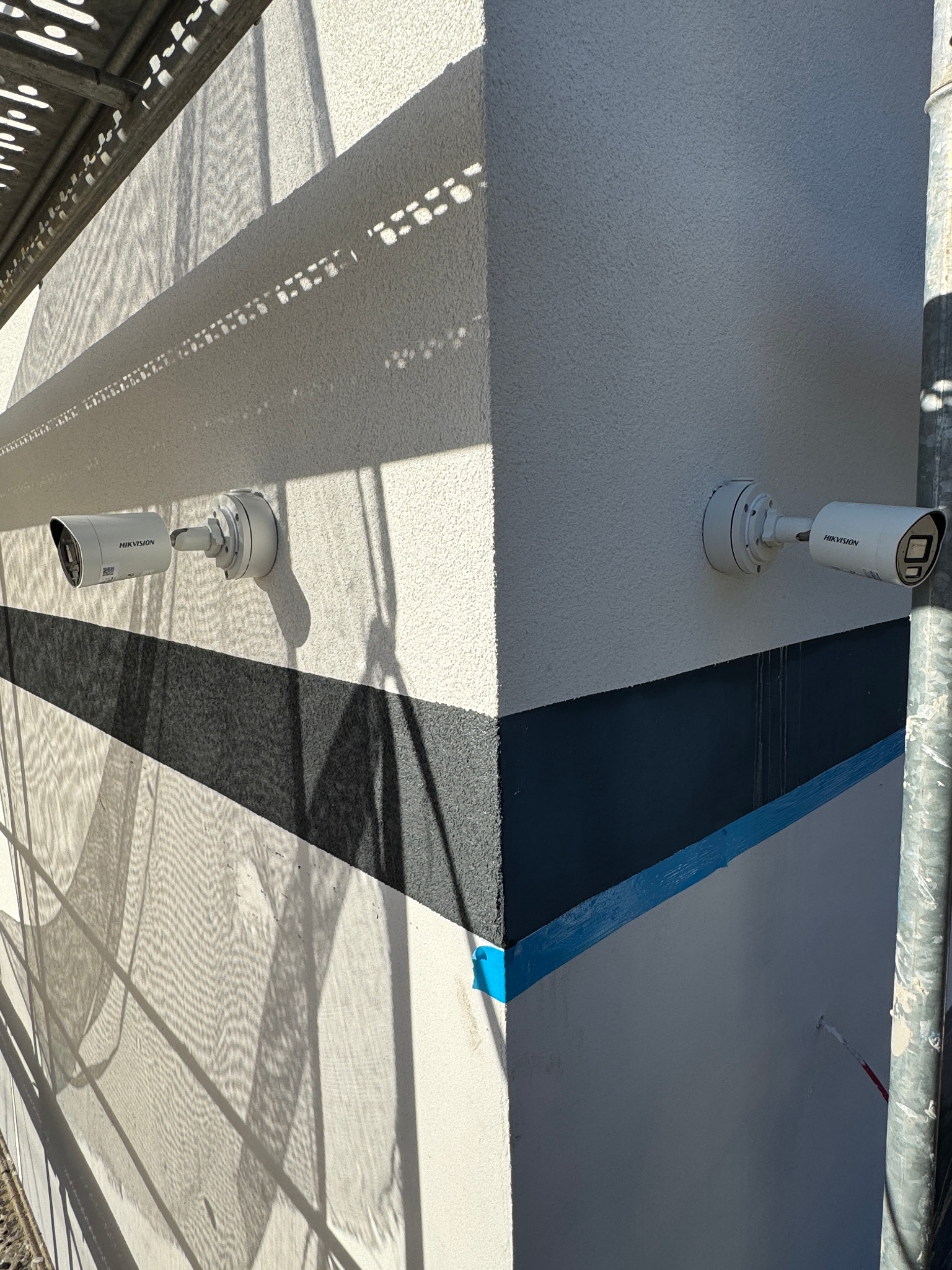 Dwie kamery monitoringu Hikvision zamontowane na świeżo pomalowanej ścianie budynku, z widocznymi śladami rusztowania i cieniem. Wykończenie elewacji w trakcie.
