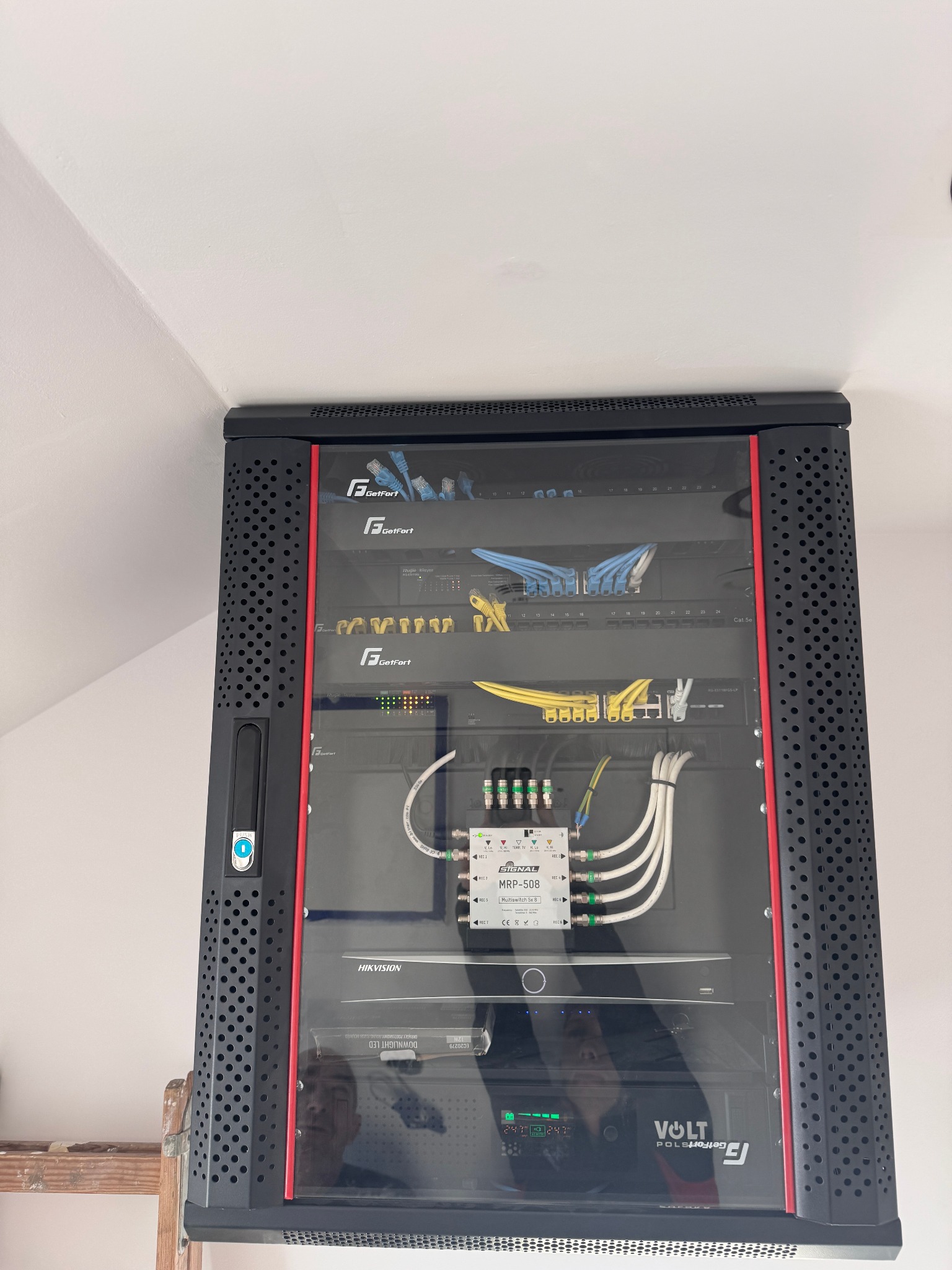Zamknięta szafa rack z zainstalowanymi urządzeniami sieciowymi i monitoringowymi, widoczne kable Ethernet i zasilające, sprzęt marek GetFort, Hikvision i Volt.