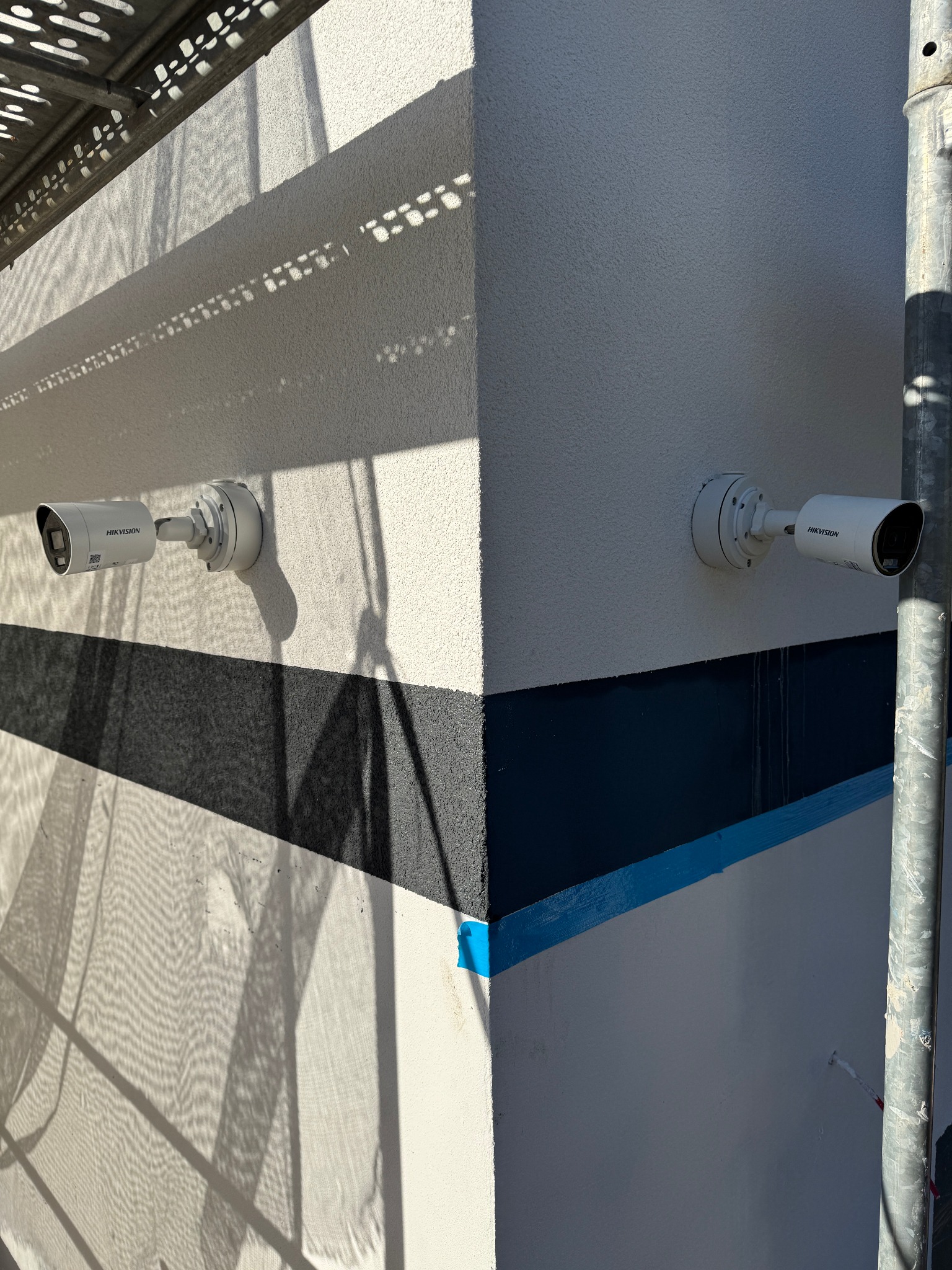 Dwie kamery monitoringu Hikvision zamontowane na elewacji budynku z widocznymi cieniami rusztowania i fragmentami malowania. Jasne światło słoneczne.