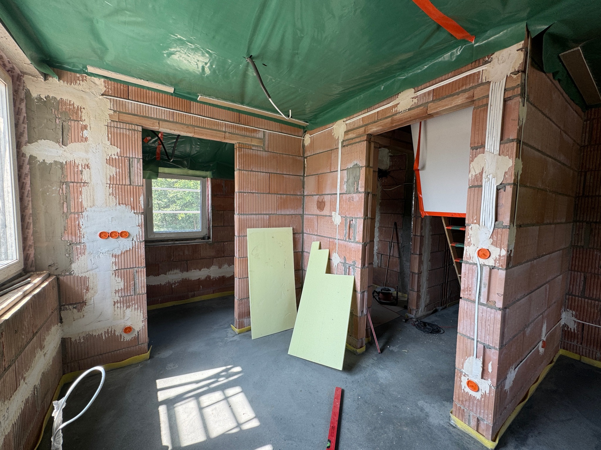 Surowy stan budynku z cegły: widoczne kable elektryczne w ścianach, okna, zielona folia na suficie. Etap instalacji elektrycznej w trakcie budowy.