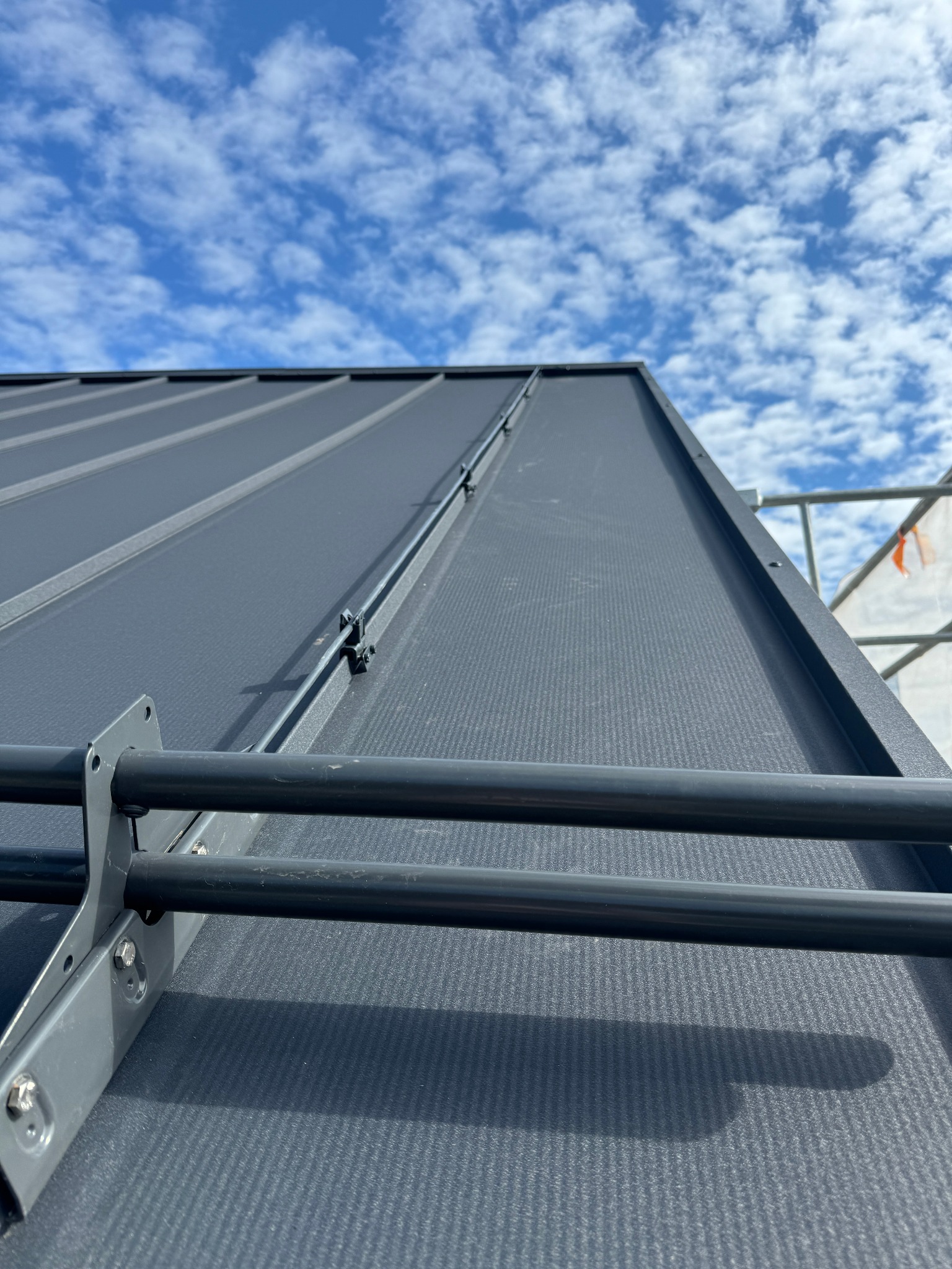 Szara blacha na dachu z zamontowaną instalacją odgromową i widocznym fragmentem rusztowania na tle błękitnego nieba z chmurami.