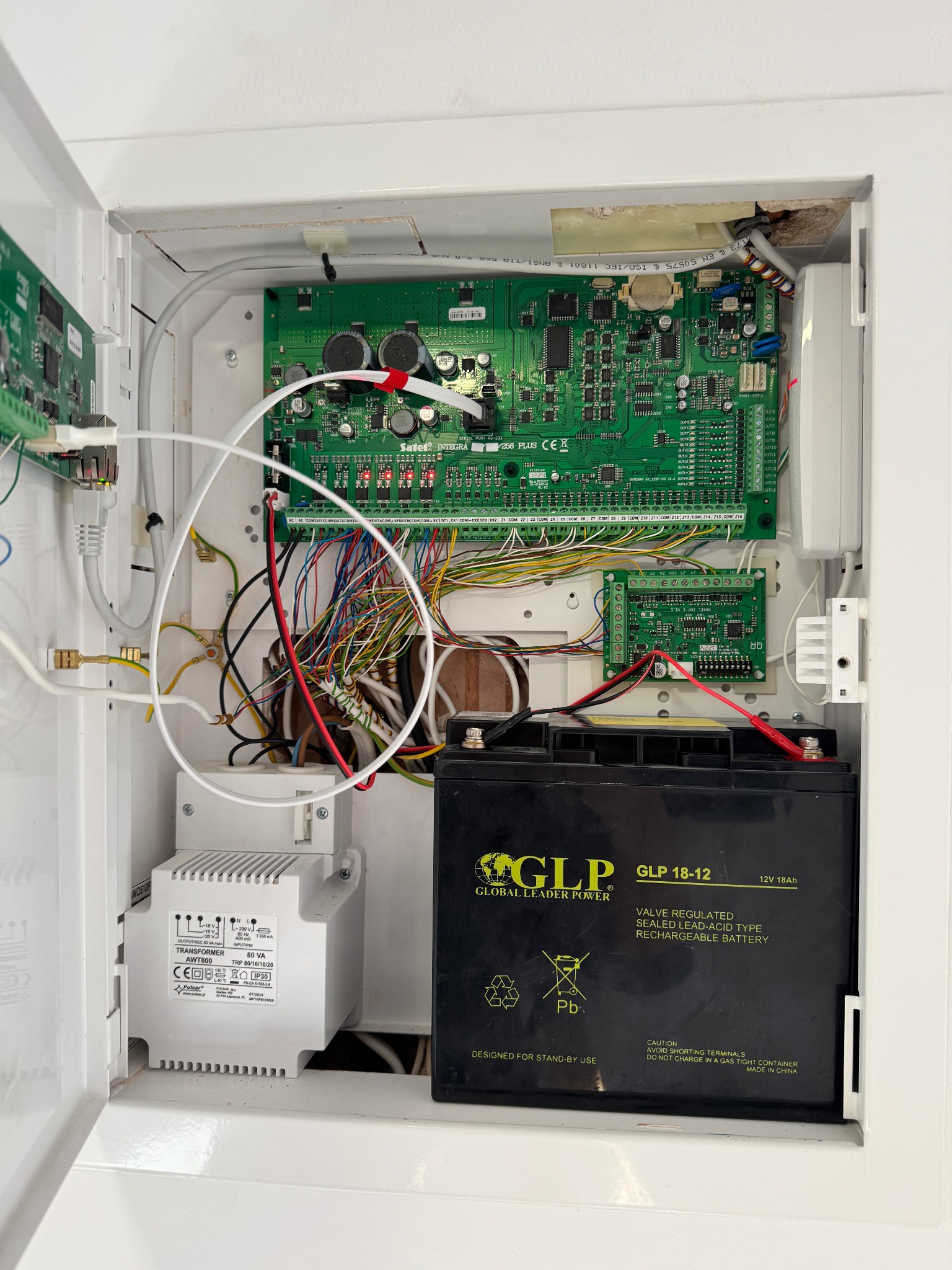 Otwarta szafka z elektroniką alarmową: płyty główne Satel Integra, transformator Pulsar AWT800 i akumulator GLP 18-12. Widoczne liczne przewody i złącza.