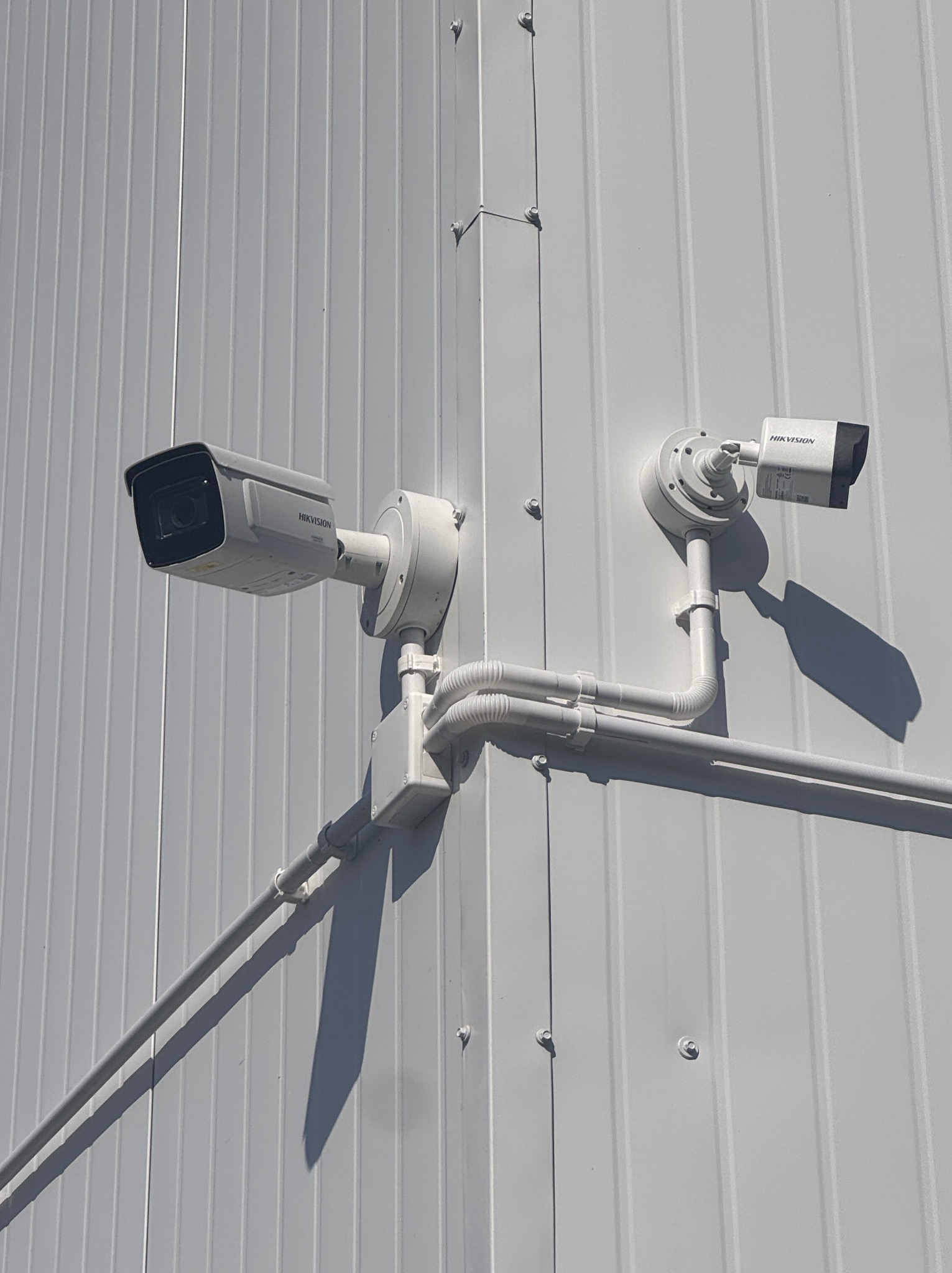 Dwie kamery Hikvision zamontowane na białej, metalowej ścianie budynku. Instalacja kabli w peszlach. Widoczne mocowania i cień kamery.
