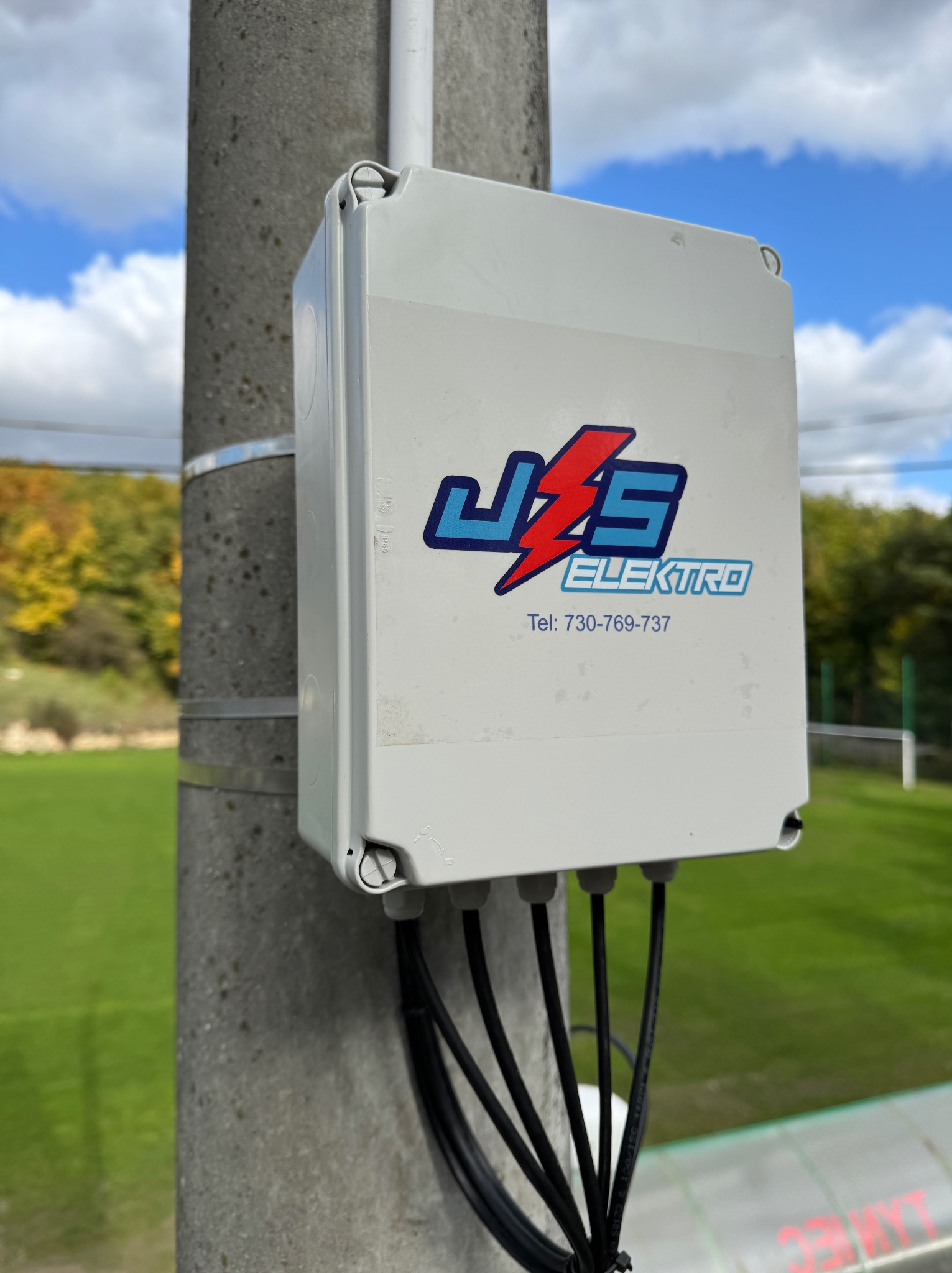 Szara skrzynka elektryczna z logo JZS Elektro zamontowana na betonowym słupie za pomocą metalowych obejm. Widoczne kable wychodzące ze skrzynki.