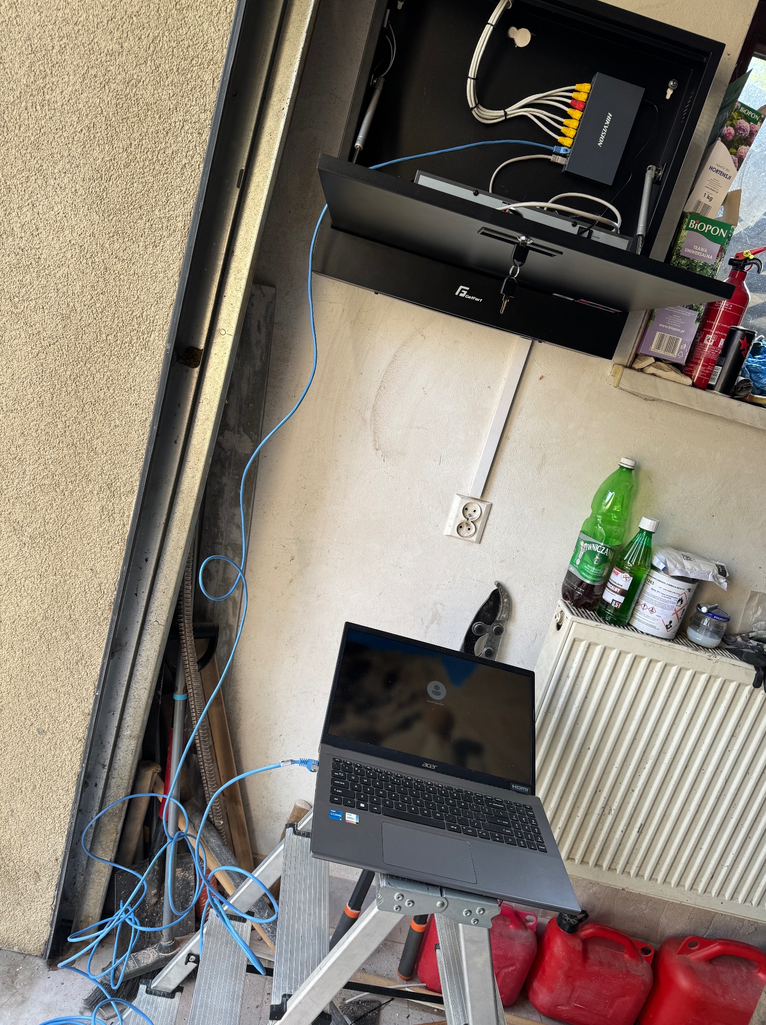 Konfiguracja systemu alarmowego: laptop podłączony do centrali alarmowej Hikvision w szafce GetFort, kable, drabina, akcesoria. Ściana z gniazdkiem i kaloryferem w tle.