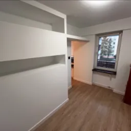 Białe ściany z wnękami i listwami przypodłogowymi po remoncie, widok na okno i fragment podłogi z paneli imitujących drewno.