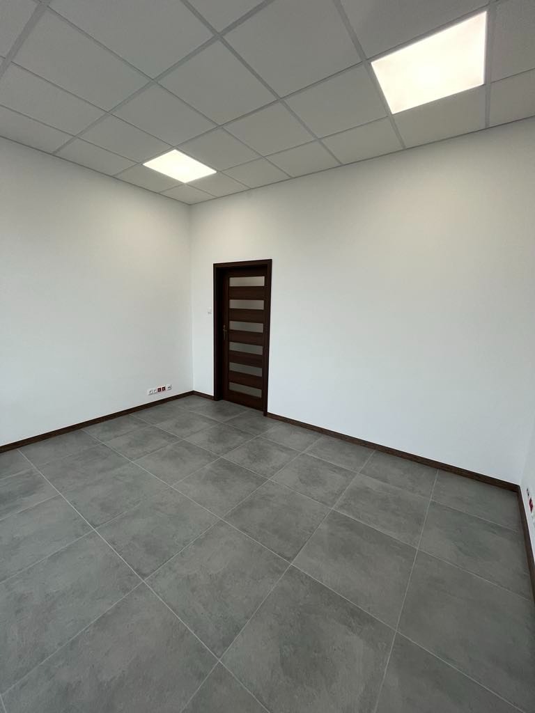 Wnętrze biura z szarymi płytkami podłogowymi, białymi ścianami i ciemnymi listwami przypodłogowymi oraz drzwiami z przeszkleniami. Sufit podwieszany z wbudowanymi panelami LED.