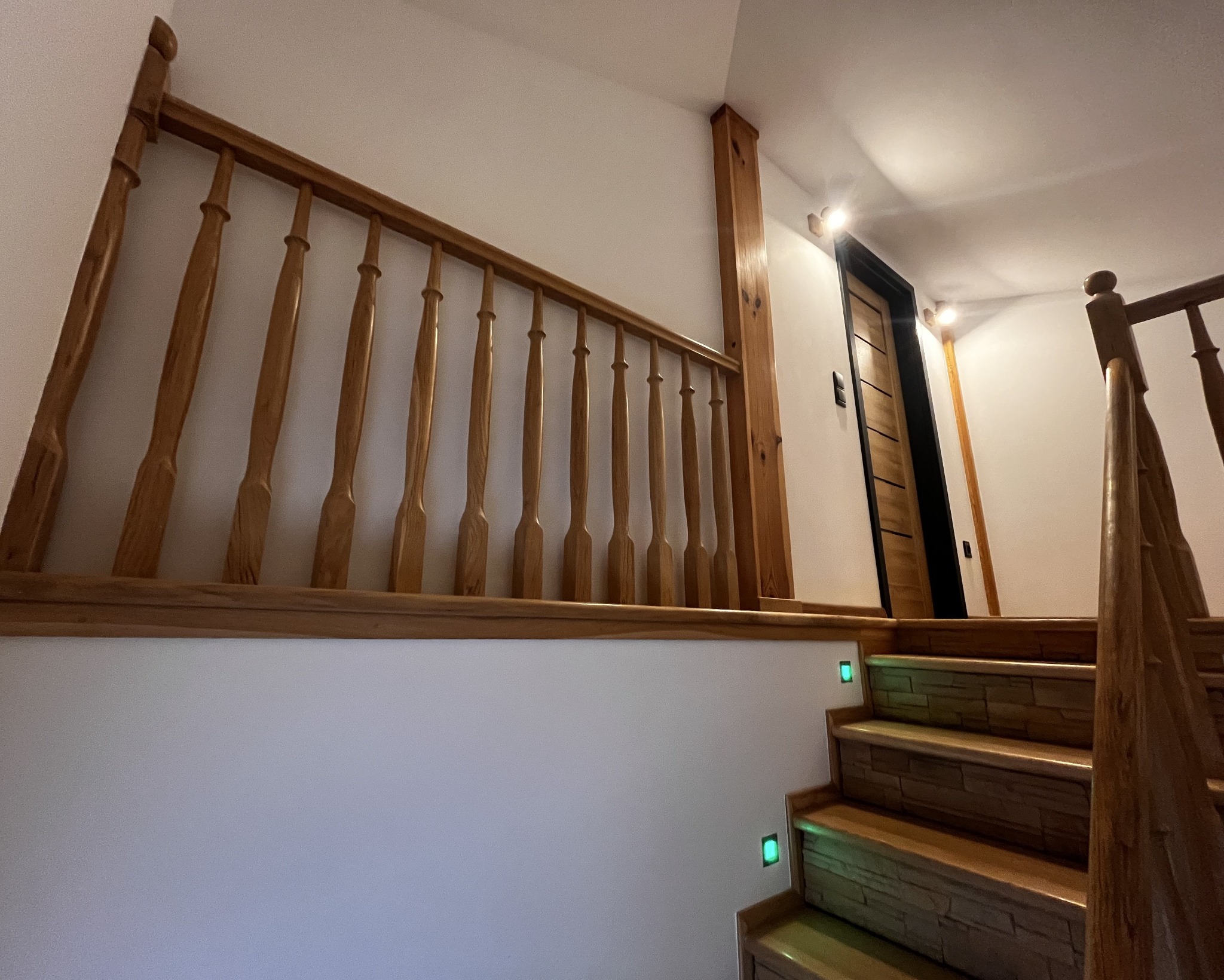 Drewniane schody z podświetlanymi stopniami i balustradą z toczonymi elementami, prowadzące do drzwi z czarną ościeżnicą.