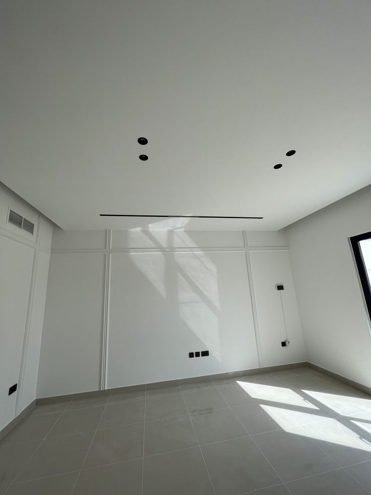 Pusty, jasny pokój z białymi ścianami z dekoracyjnymi listwami, szarą podłogą z płytek, czarnymi punktowymi lampami sufitowymi i poziomym czarnym panelem wentylacyjnym