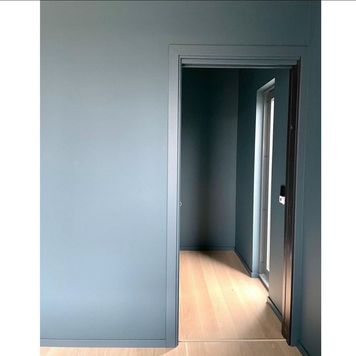 Wnętrze po malowaniu na jednolity, pastelowy odcień błękitu, widoczne ściany, framuga drzwi i parkiet z jasnego drewna.