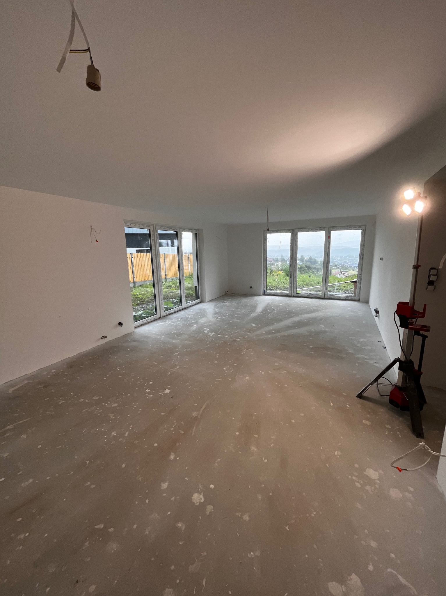 Wnętrze pokoju w stanie deweloperskim z widocznymi śladami po gładzi na podłodze, surowymi ścianami i niezabezpieczonym kablem oświetleniowym, oświetlone lampą budowlaną.