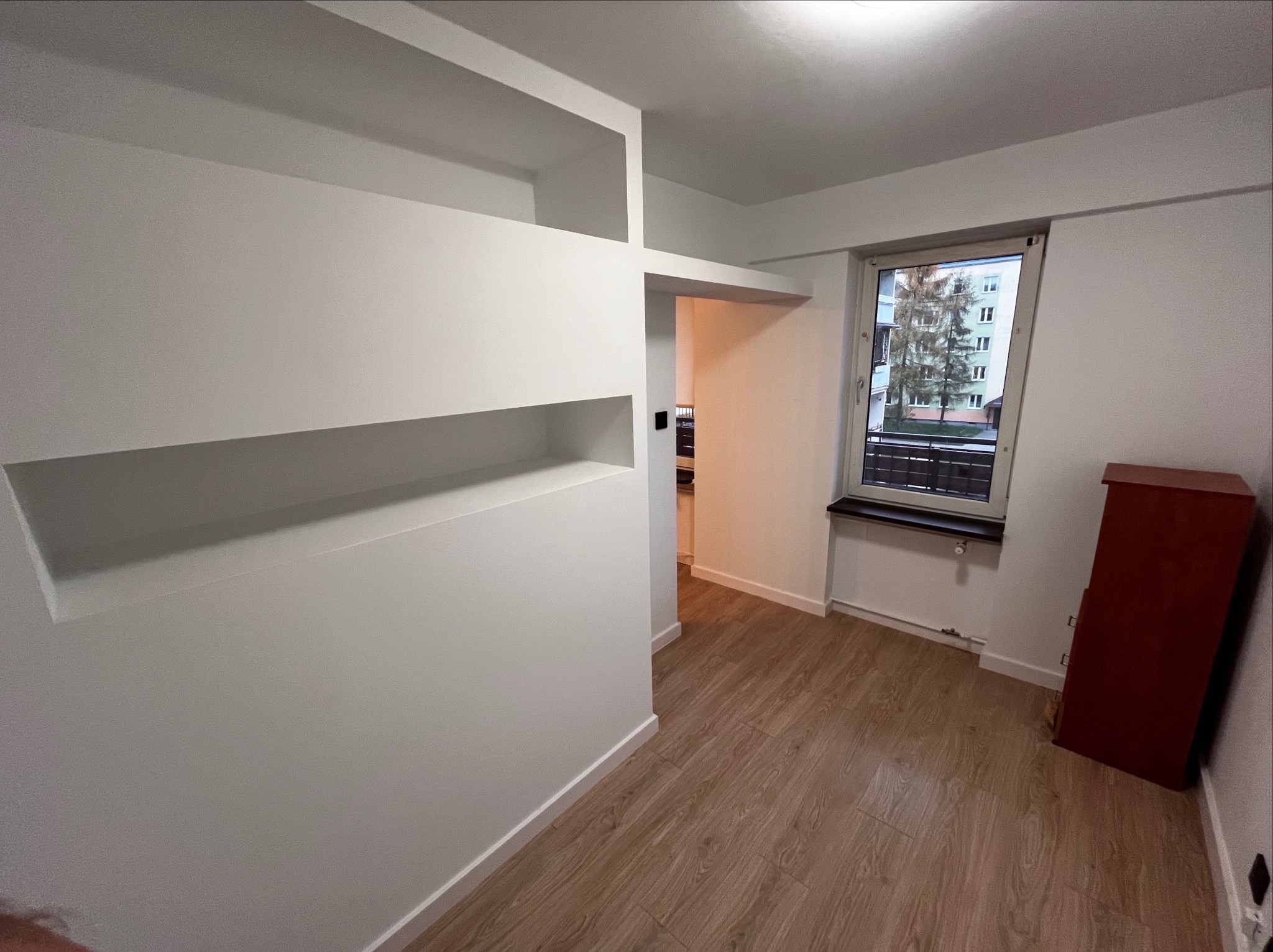 Białe ściany z wnękami i listwami przypodłogowymi po remoncie, widok na okno i fragment podłogi z paneli imitujących drewno.