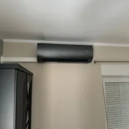 Czarny klimatyzator Sinclair zamontowany na ścianie w jasnym pokoju, obok okna z żaluzjami i szafki. Widoczny wyświetlacz temperatury.