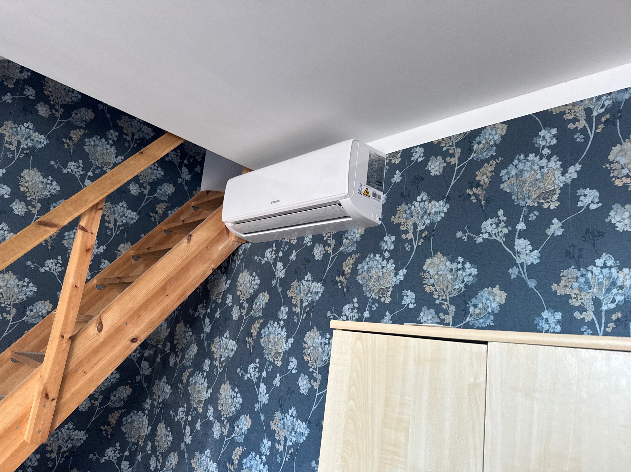 Nowoczesny klimatyzator ścienny Sencor w pokoju z tapetą w kwiaty i drewnianymi schodami. Widok z dołu, oświetlenie naturalne.