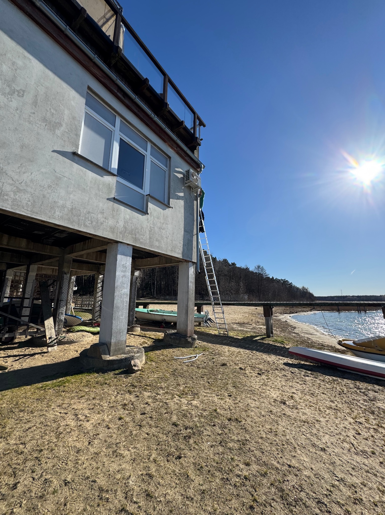 Montaż klimatyzacji na elewacji budynku z balkonem, widok z dołu, drabina oparta o ścianę, w tle jezioro i plaża. Słoneczny dzień.