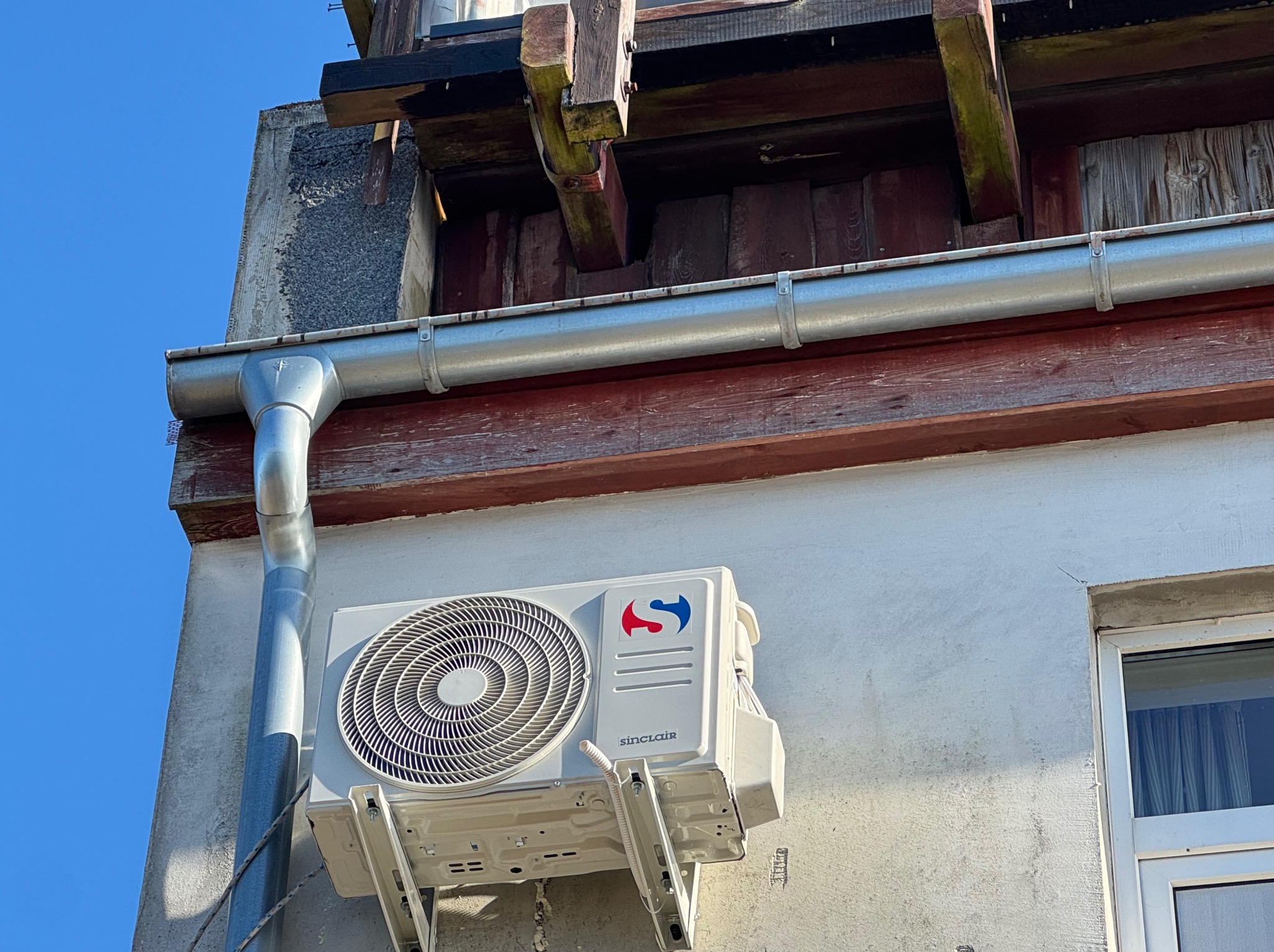 Biała klimatyzacja Sinclair zamontowana na elewacji budynku obok rynny, z fragmentem okna i niebieskim niebem w tle.