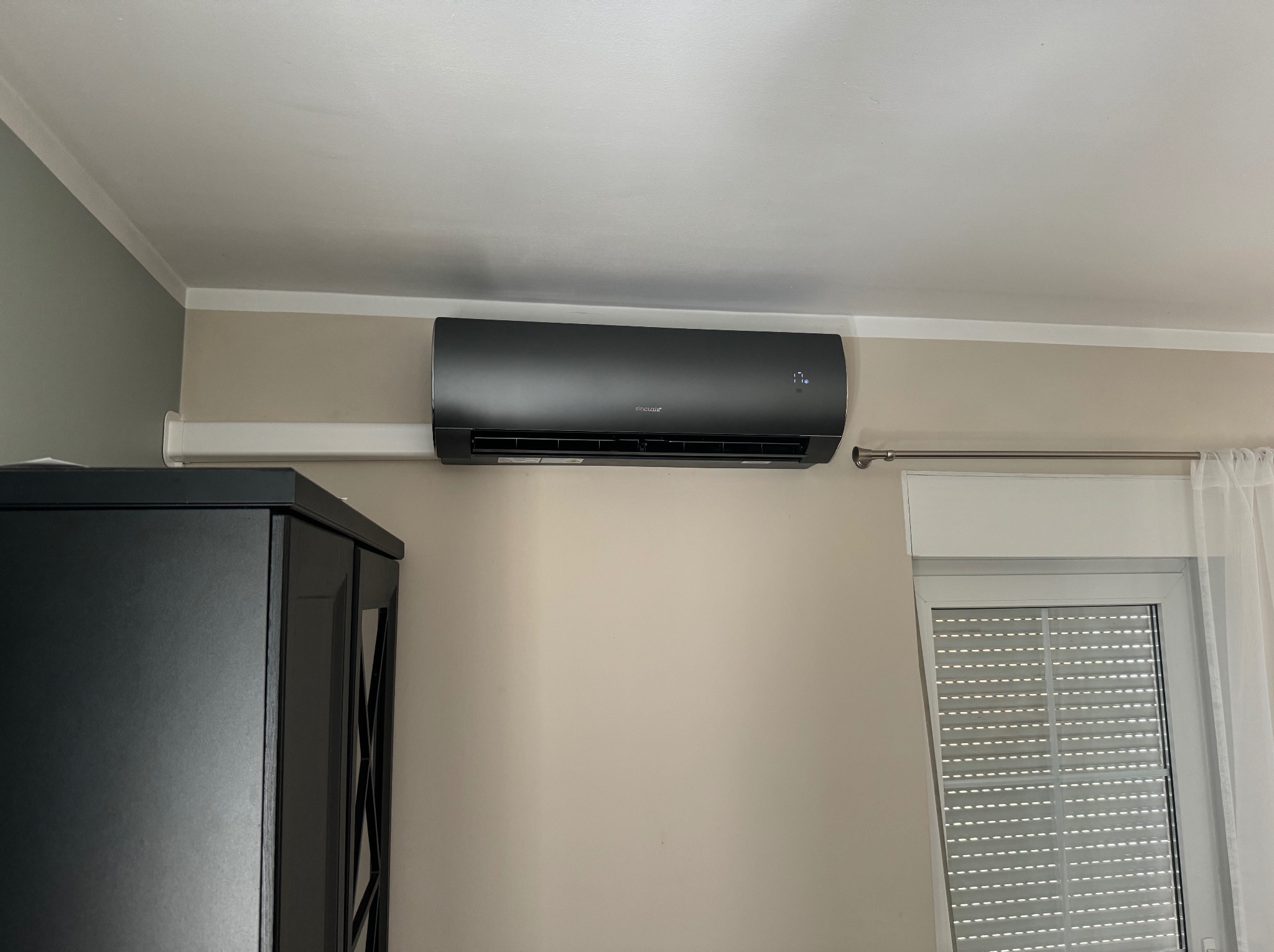 Czarny klimatyzator Sinclair zamontowany na ścianie w jasnym pokoju, obok okna z żaluzjami i szafki. Widoczny wyświetlacz temperatury.