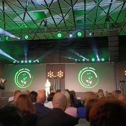 Dj.Marciz - Konferencja z prelegentem na scenie oświetlonej zielonymi światłami i reflektorami, widoczny tłum słuchaczy oraz operator kamery.