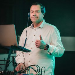 Dj.Marciz - Mężczyzna w słuchawkach daje kciuk w górę, stoi za konsoletą DJ-ską z laptopem, oświetlony zielonym światłem.